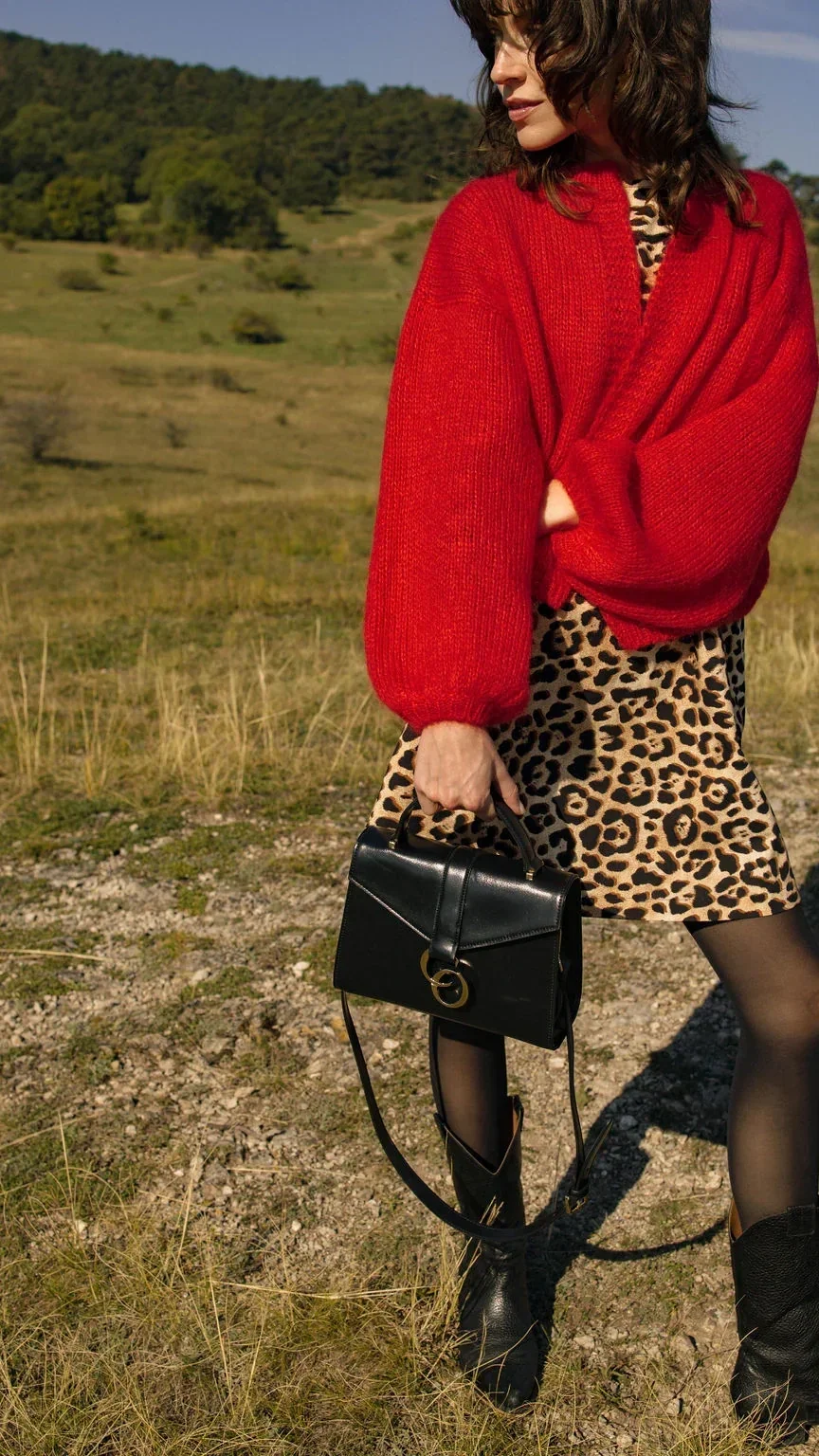 Frau trägt eine rote Strickjacke, ein leo-muster Kleid, schwarze Strumpfhosen, schwarze Stiefel und hält eine schwarze Handtasche in der Hand. Sie steht in einer offenen, grünen Landschaft.