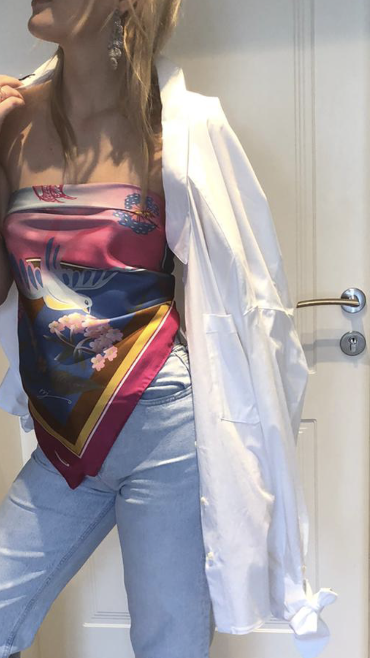 Eine Person trägt einen bunten, bedruckten Bandeau-Top, eine weiße Jacke, hellblaue Jeans, und große silberne Ohrringe, steht vor einer weißen Tür.