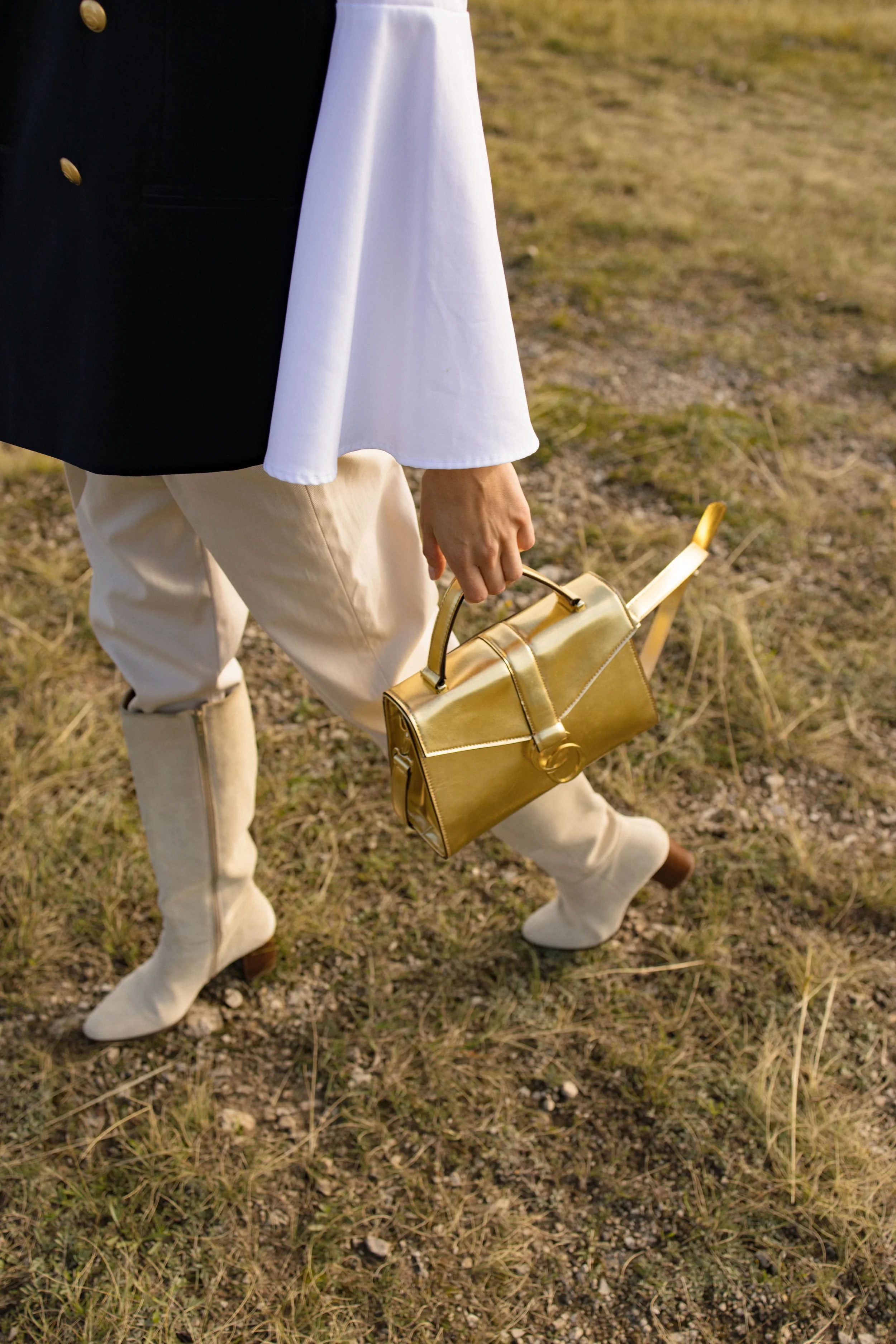 Nahaufnahme einer Person, die violette Stiefel, beige Hosen, eine weiße Bluse und einen schwarzen Mantel trägt, und eine goldene Handtasche hält, während sie auf einem Feld steht.