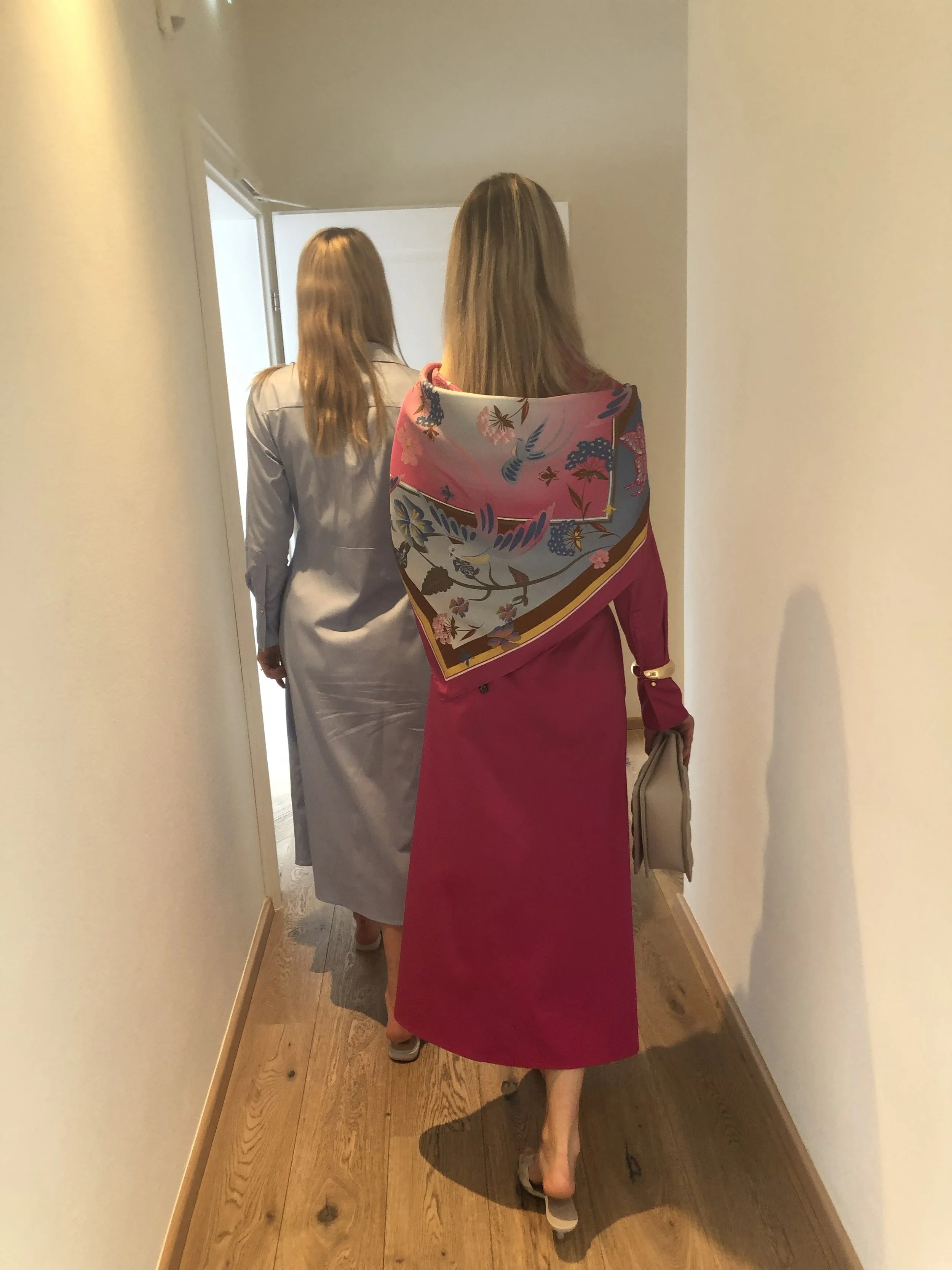 Zwei Frauen gehen den Flur eines Hauses entlang, eine trägt ein pinkes Kleid mit einen bunten Schal, die andere trägt ein graues Kleid.