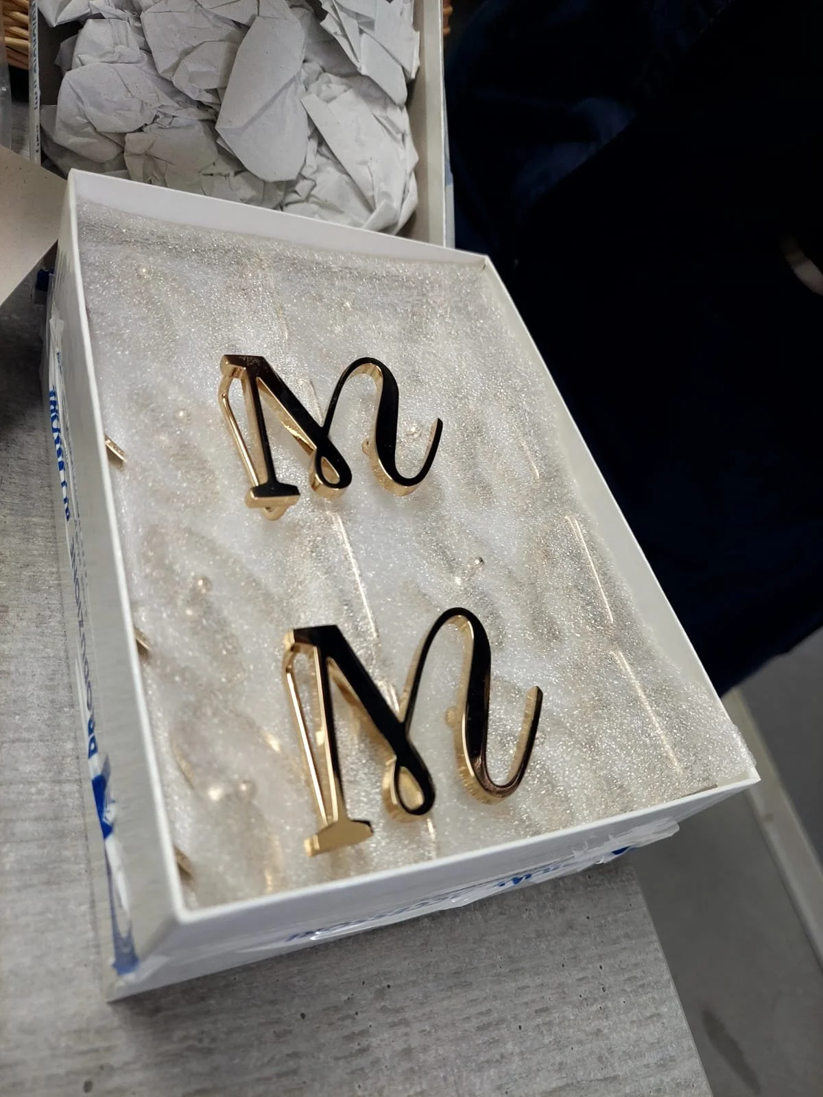 Eine offene Verpackung mit zwei dekorativen Buchstaben M und N, die auf einer weißen, glitzernden Oberfläche liegen.