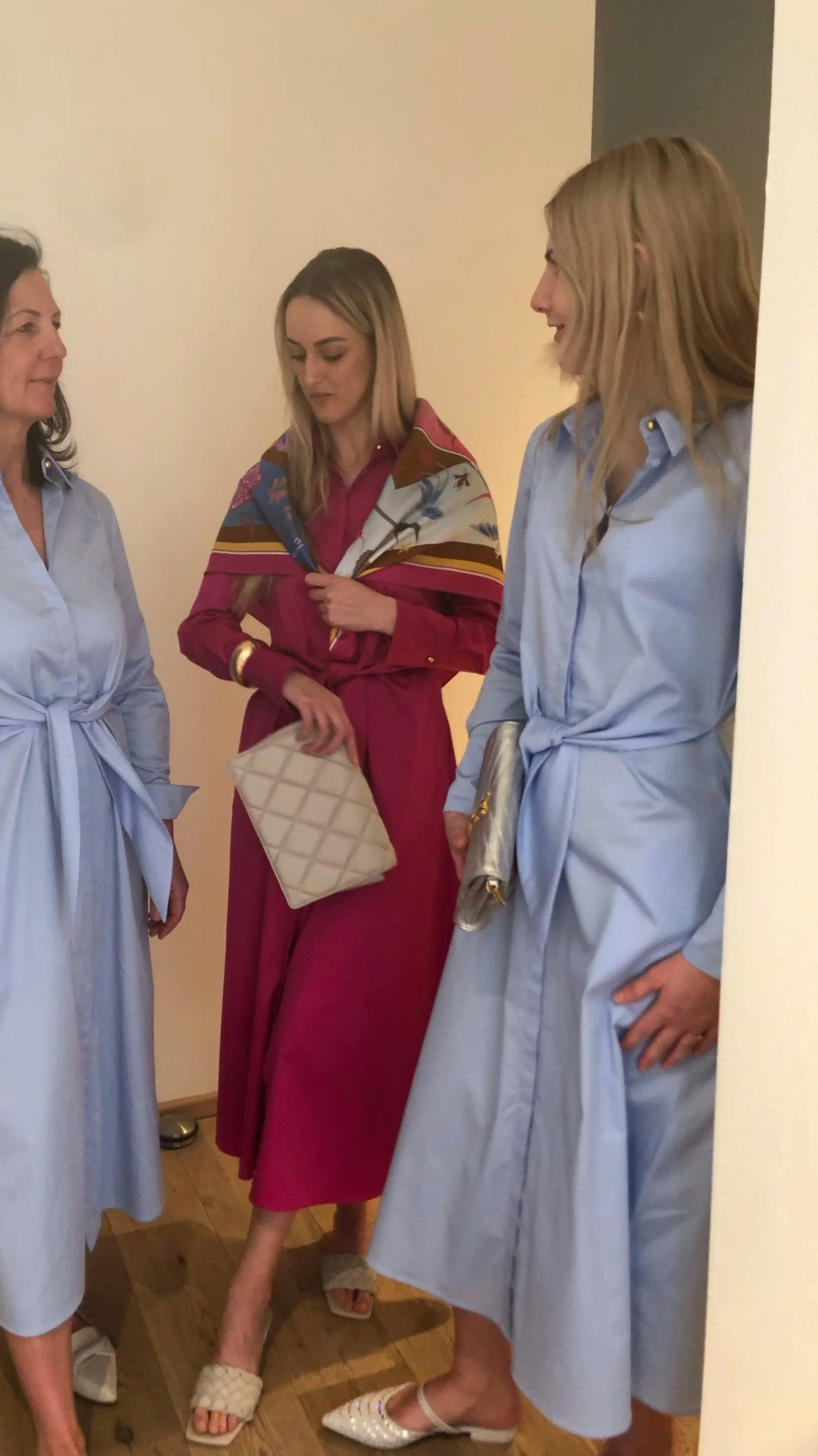 Drei Frauen stehen im Gespräch, zwei tragen hellblaue Kleider und eine trägt ein pinkfarbenes Kleid mit Schal. Alle halten Handtaschen.