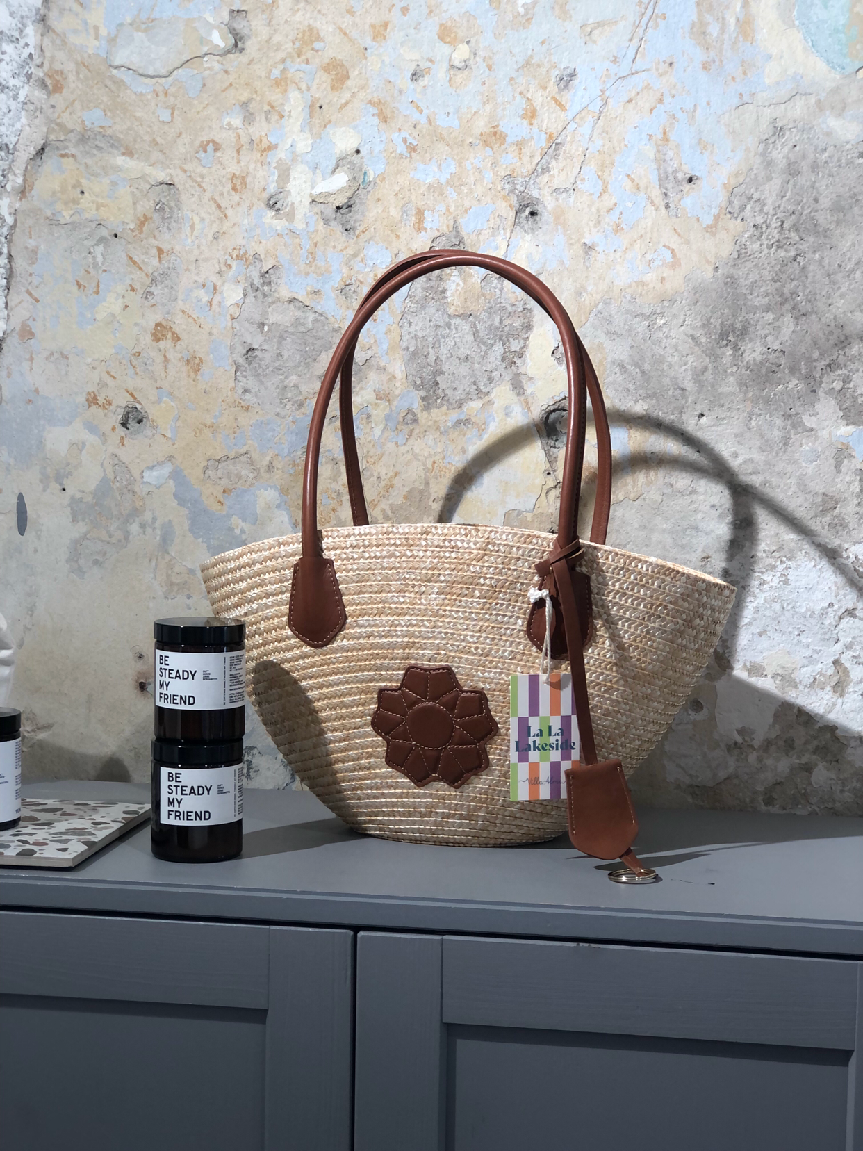 Strohkorb-Tasche mit braunen Ledergriffen und Applikation, auf einem grauen Möbelstück gegen eine alte, abblätternde Wand. Daneben befinden sich zwei schwarze Dosen mit weißem Etikett.