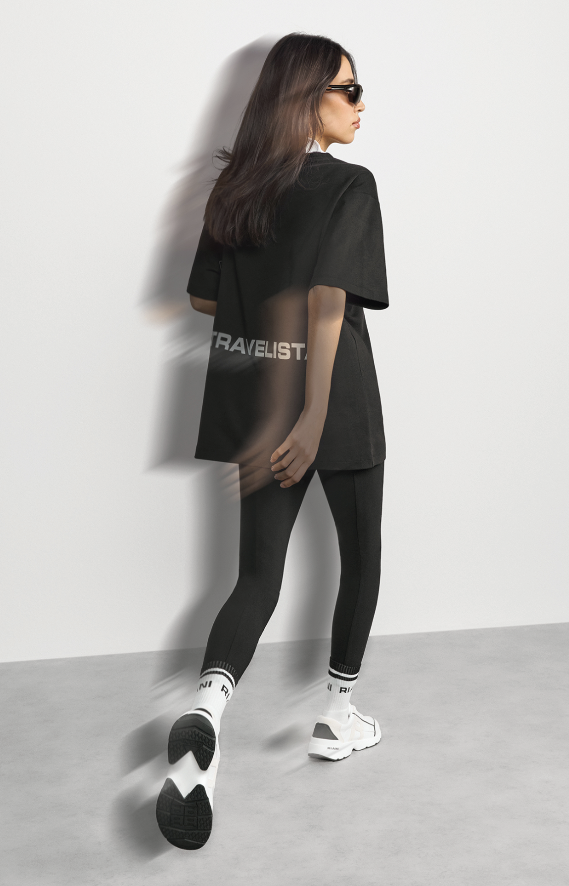 Junge Frau in schwarzer Sportkleidung, weißer sportlicher Schuhe, schwarze Sonnenbrille, vor weißer Wand, Schatten im Hintergrund, sportlicher Look, moderner Style.