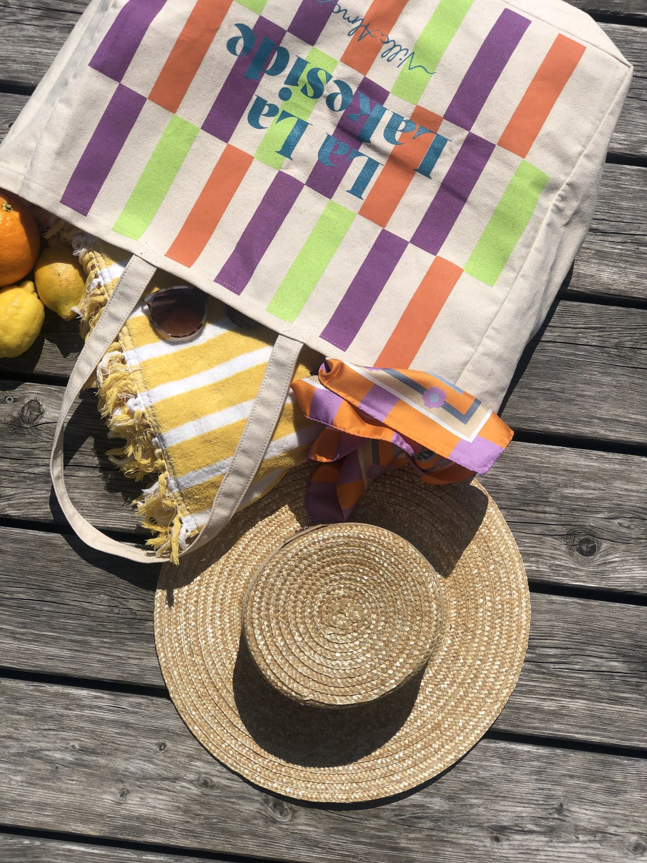 Strandtasche mit bunten Streifen und Text, Sonnenbrille, bunter Schal, Sonnenhut, Früchte, auf Holzbohlen. Ideal für einen Tag am Strand oder im Park.