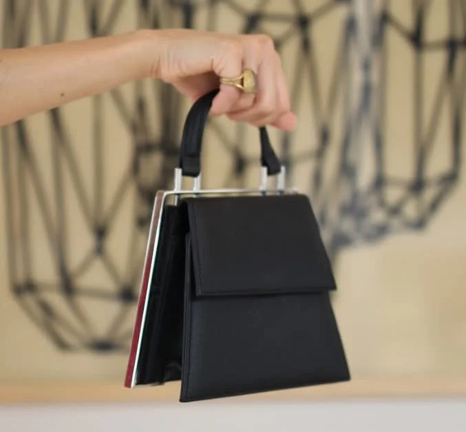 Hand hält eine schwarze Handtasche mit mehreren Büchern darin. Hintergrund mit geometrischem Wanddekor.