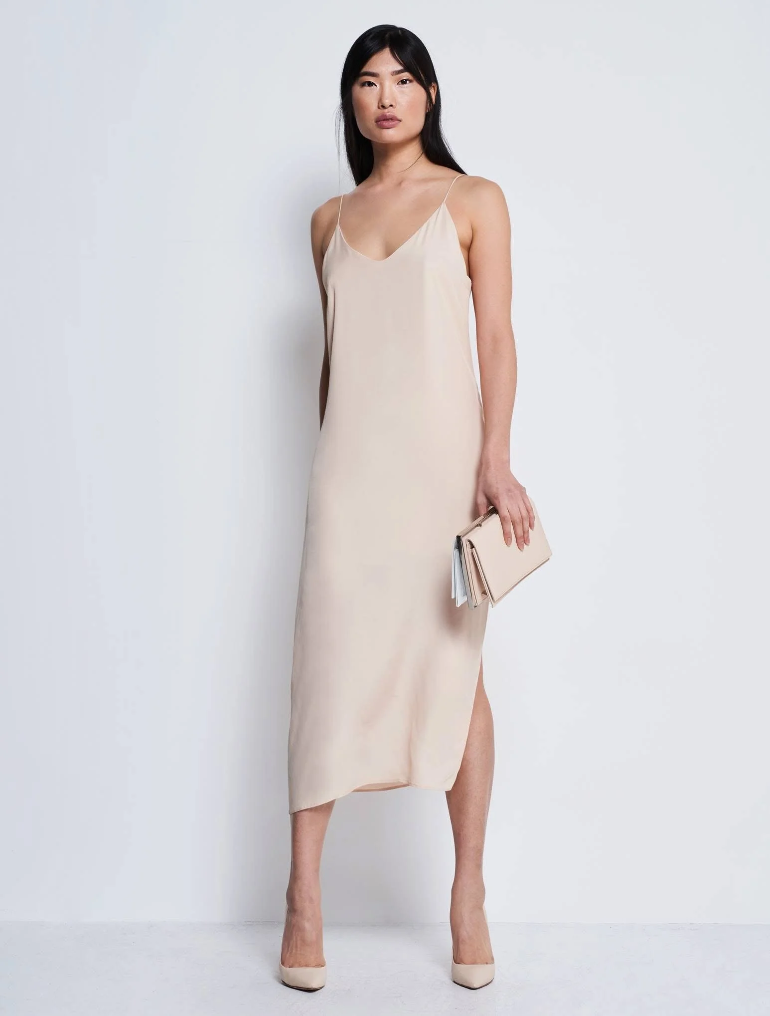 Frau in beigem, schulterfreiem Kleid mit Spaghettiträgern, die eine Clutch hält und auf hellen High Heels steht.