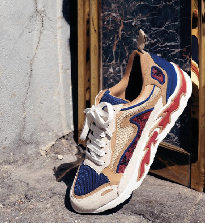 Farbenfroher Turnschuh mit beige, blau, rot und schwarzen Details, auf einem grauen Boden vor einer Wand mit Marmor- und Holzdekoration.