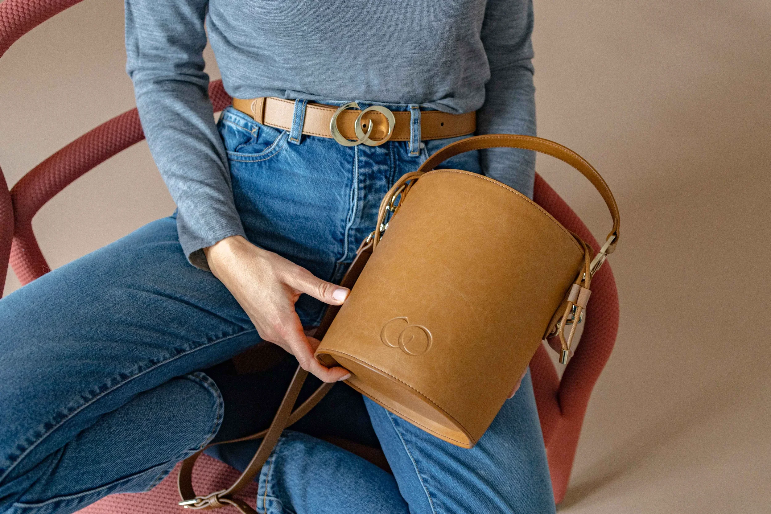 Person sitzt in einem roten Stuhl, trägt eine graue Jacke, blaue Jeans und einen beige Gürtel mit einem goldfarbenen Schnallen. Die Person hält eine braune Ledertasche.