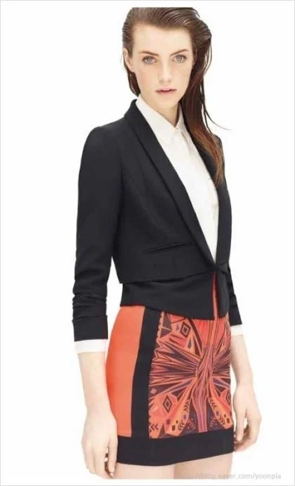 Junge Frau in formellem Outfit, schwarze Jacke und bunter Rock vor weißem Hintergrund.