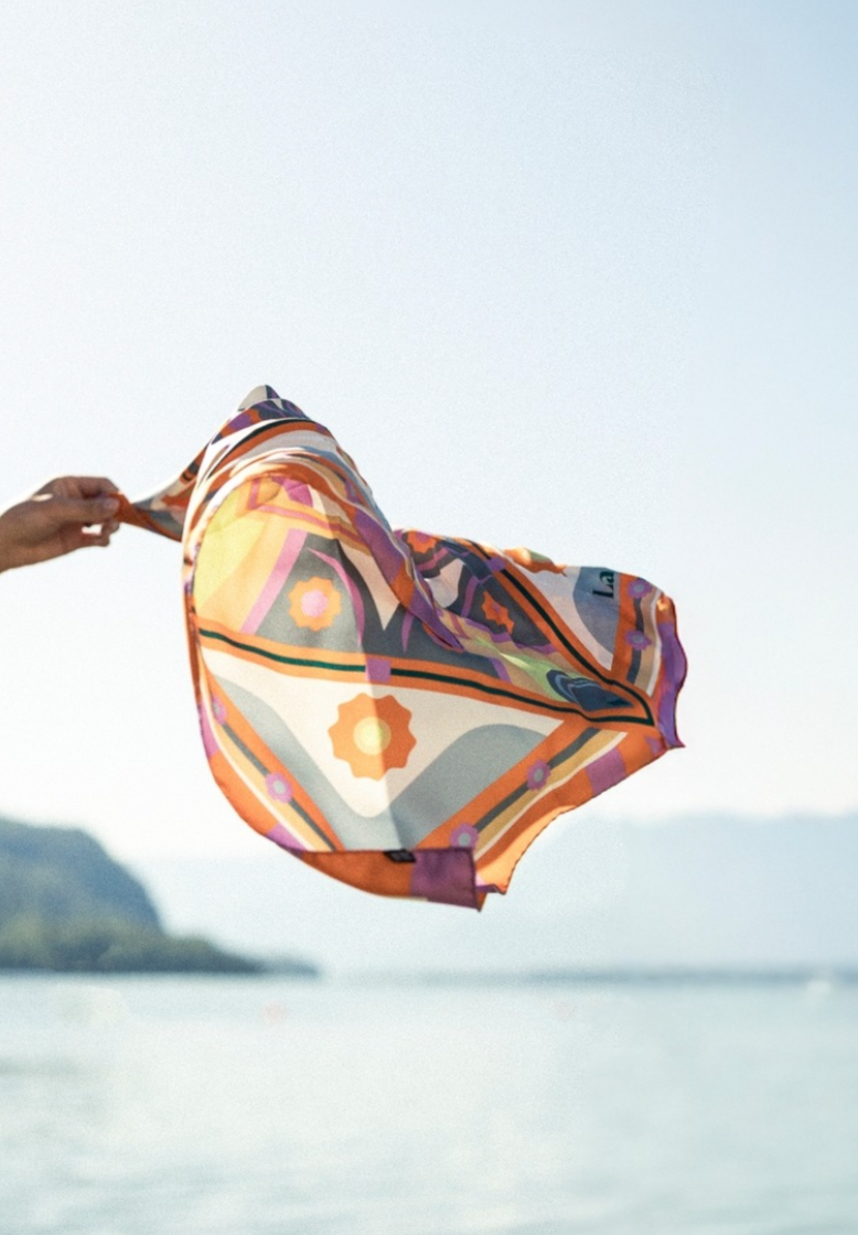 Eine Hand hält ein bunt gemustertes Tuch, das im Wind weht, vor einem Wasser- und Landschaftshintergrund.
