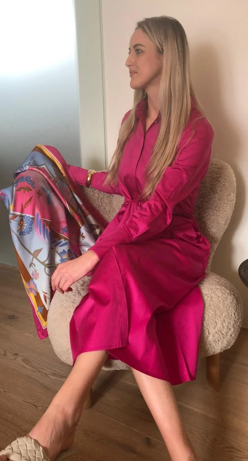 Eine Frau sitzt auf einem beige Sofastuhl, trägt ein pinkes Kleid und hält ein buntes Tuch in der Hand. Sie hat lang blondes Haar und blickt nach rechts.