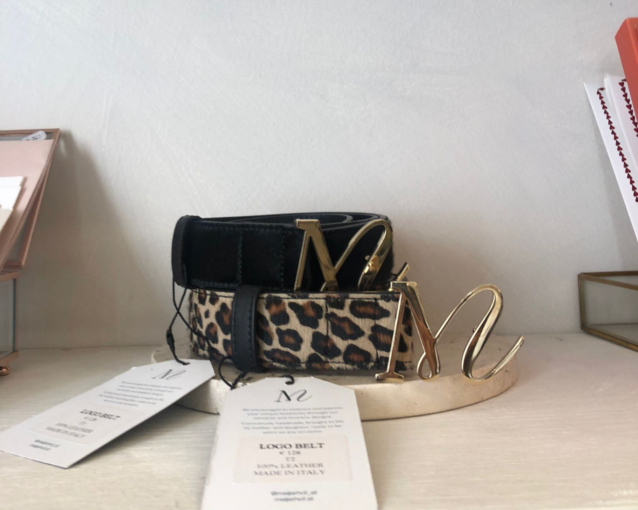 Gürtel mit goldenen Buchstaben 'M' und 'L', eines mit Leopardenmuster, auf einem Tisch mit Tags.