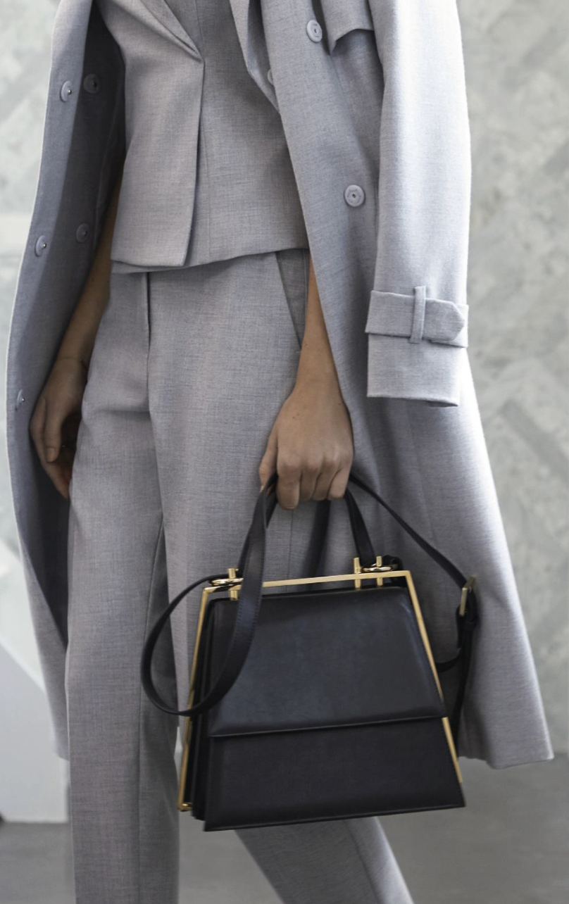 Eine Person trägt einen grauen Hosenanzug und einen passenden Mantel, hält eine schwarze Handtasche mit goldenen Akzenten.