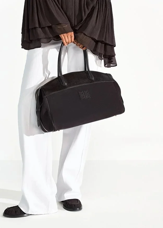 Person hält eine große schwarze Tasche in der Hand, trägt eine schwarze, rüschenartige Bluse und weiße weite Hosen, vor weißem Hintergrund.