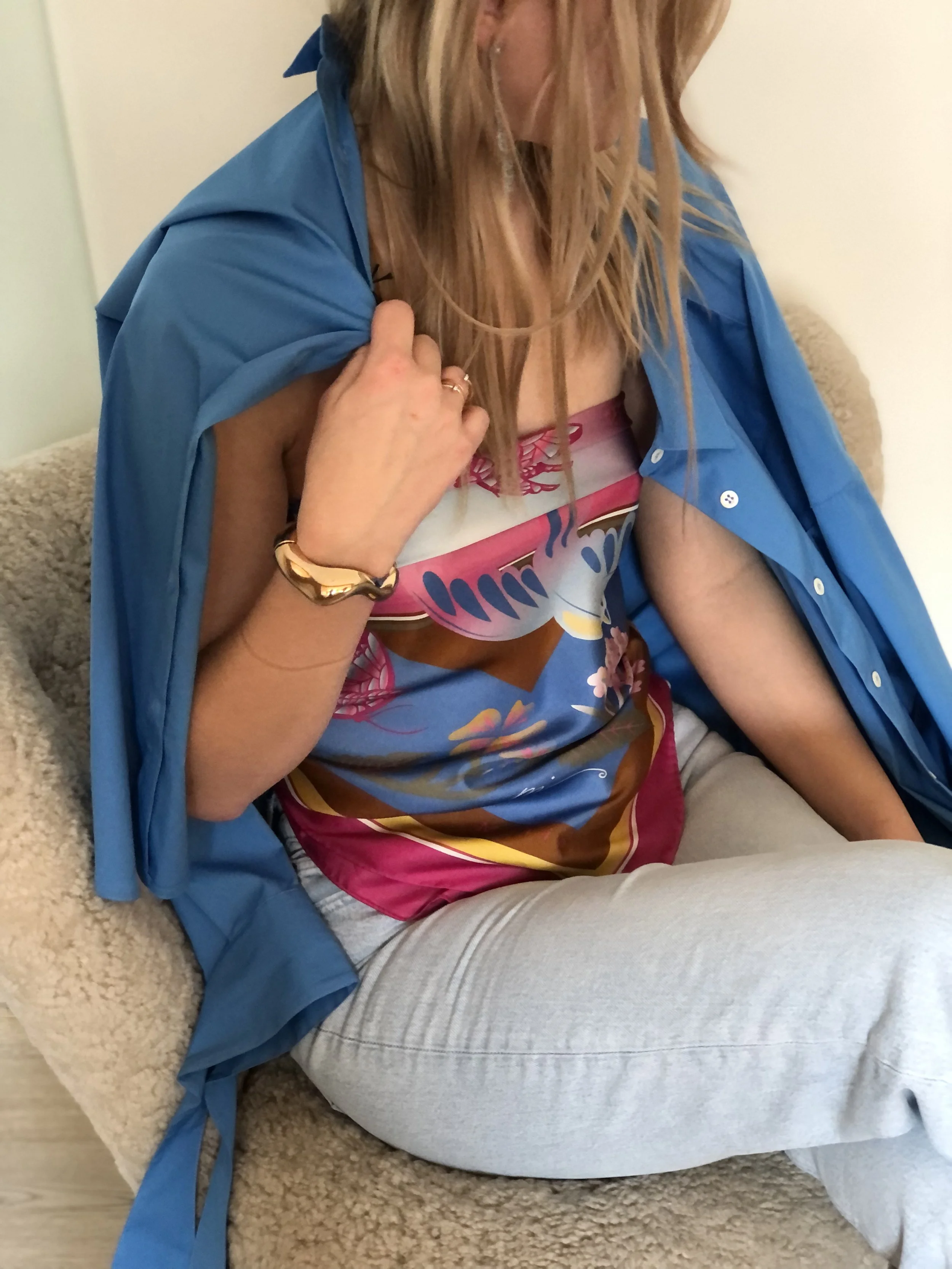 Nahaufnahme einer sitzenden Frau mit blauer Jacke, bunter Top und hellgrauen Hosen, die auf einem beige Stoffsessel sitzt.