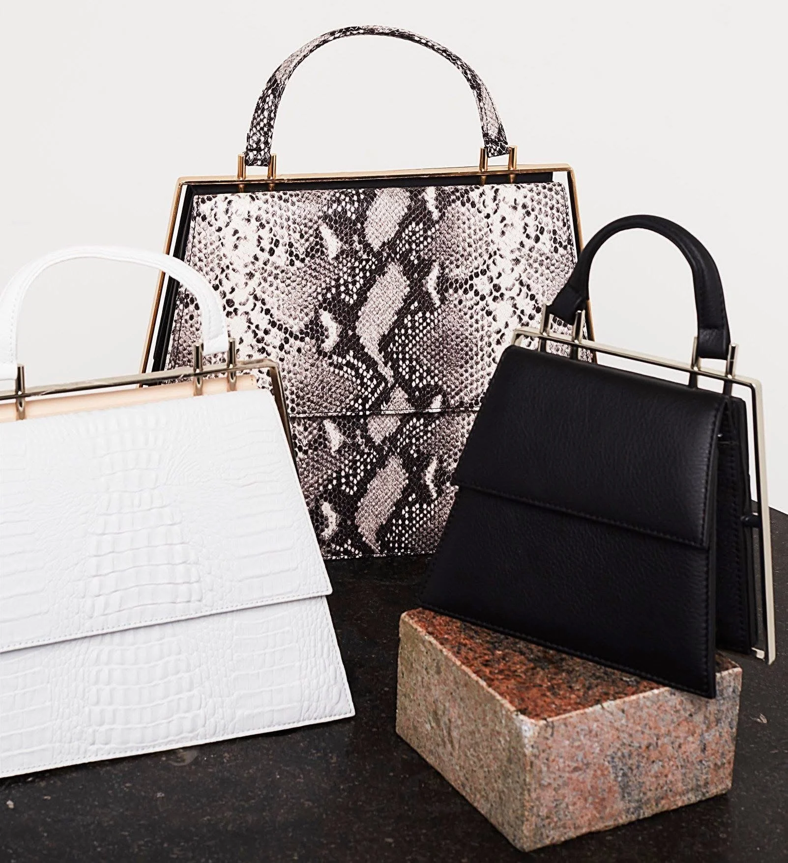 Drei Handtaschen in Schwarz, Weiß und Snake-Print, auf einer schwarzen Oberfläche mit einem Stein als Unterlage. Designed by Elisabeth Reinhartshuber
