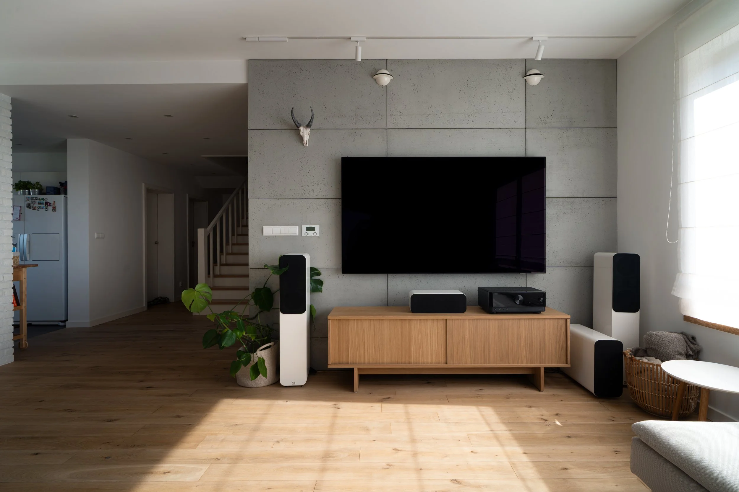 Living room with a wall-mounted flat-screen TV, wooden media console, tall white speakers, indoor plant, wall-mounted deer skull, and a window with white curtains.