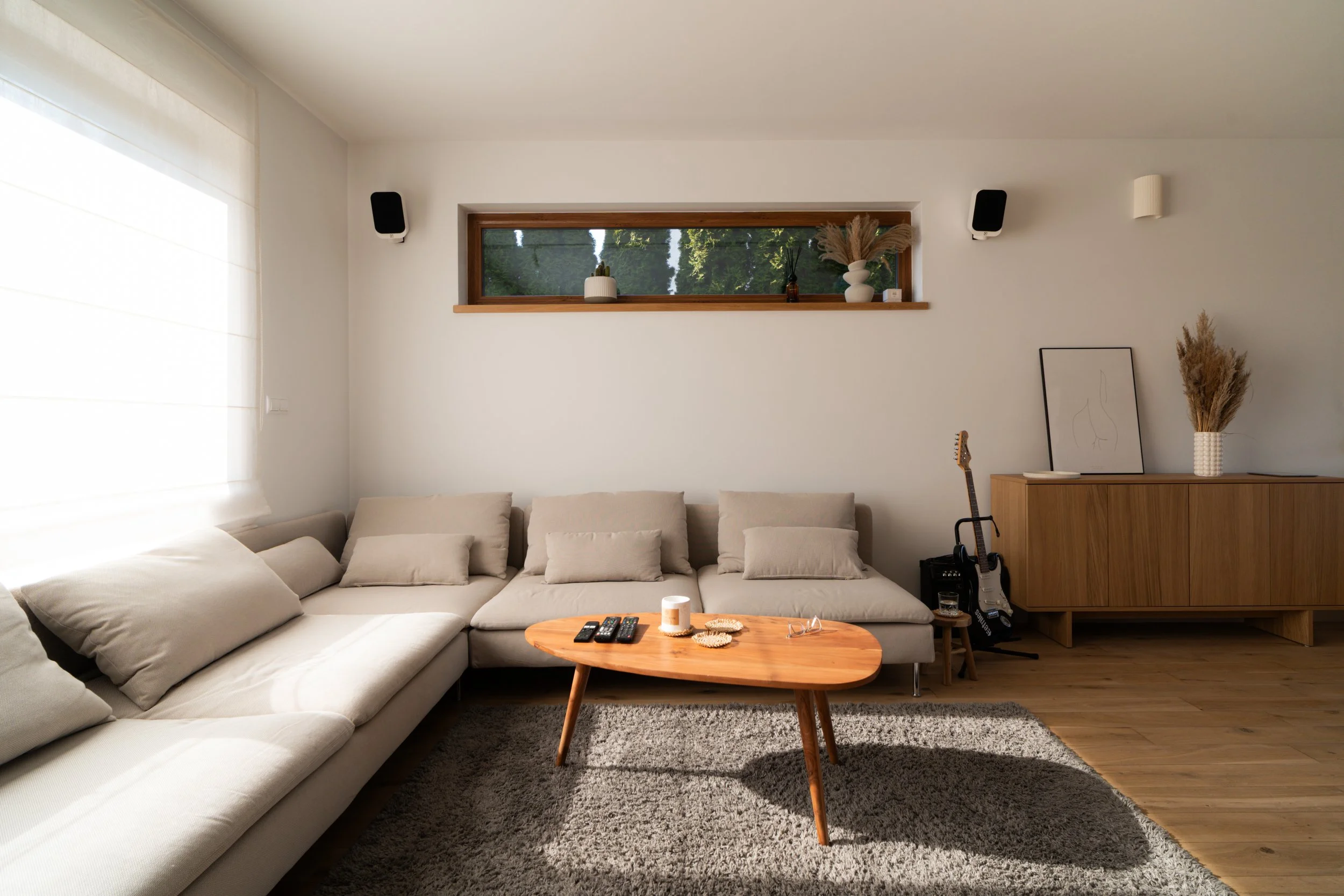 A modern living room with beige sectional sofa, wooden coffee table, and a gray area rug. The room has white walls, a long horizontal window with decorative plants, and a wooden sideboard with framed artwork, vases with dried plants, and a guitar lea