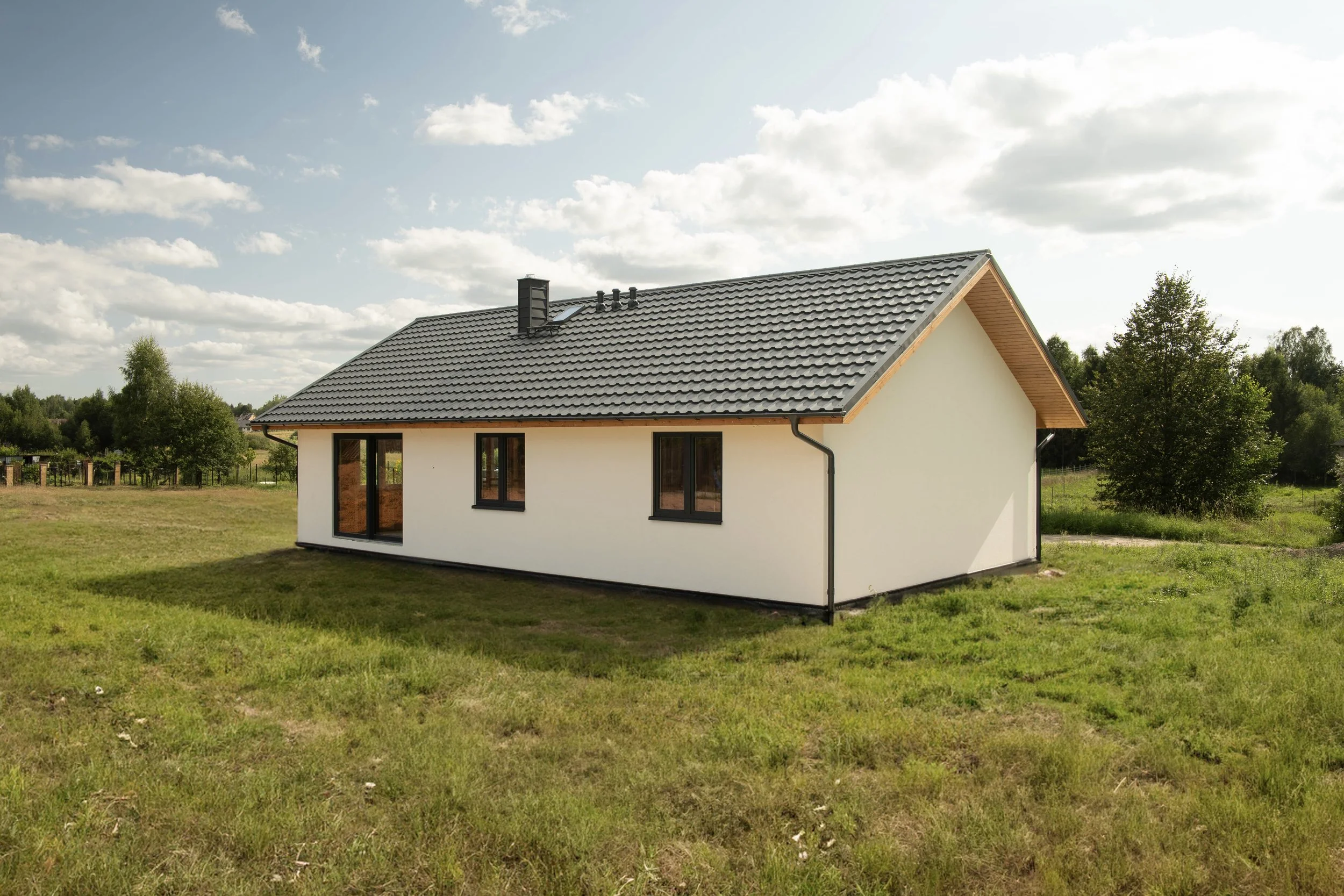 A modern single-story house with white walls, dark window frames, and a gray tiled gable roof, situated on a grassy field with trees in the background under a partly cloudy sky.
