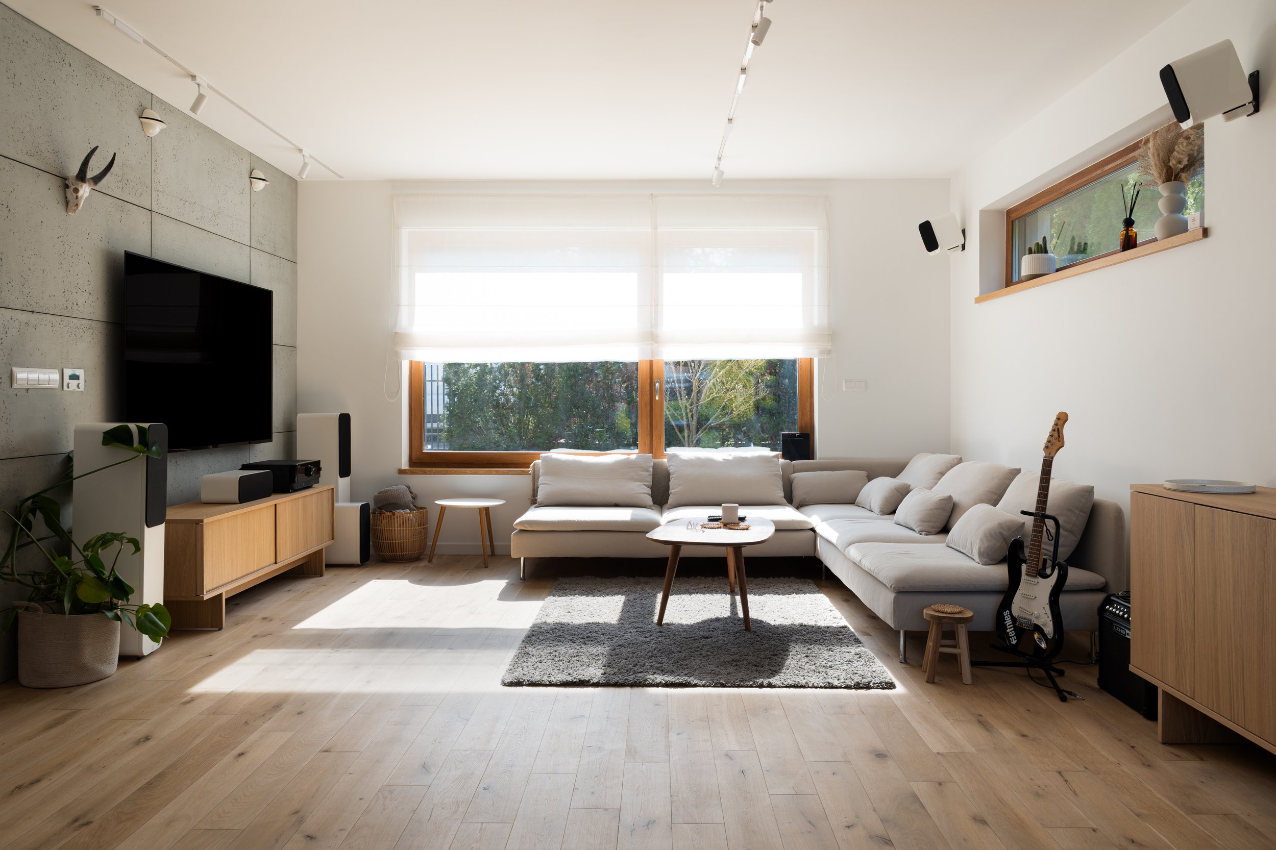 Living room with large white sectional sofa, wooden coffee table, black guitar, TV on wooden stand, large window, houseplants, and minimal decor.
