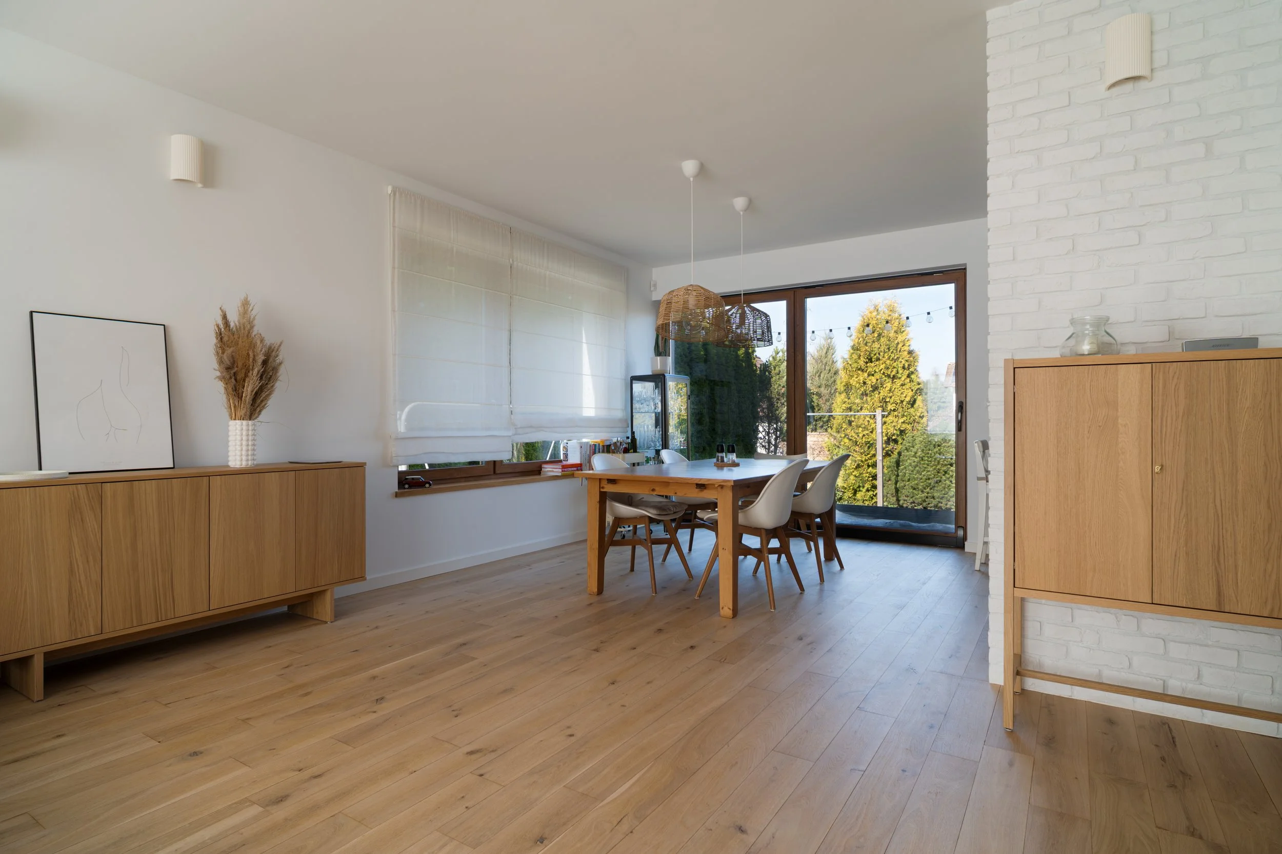 Bright dining room with wooden floor, a wood dining table with white chairs, large windows, a sliding glass door with a balcony, wooden sideboards, minimalist decor, and natural light.