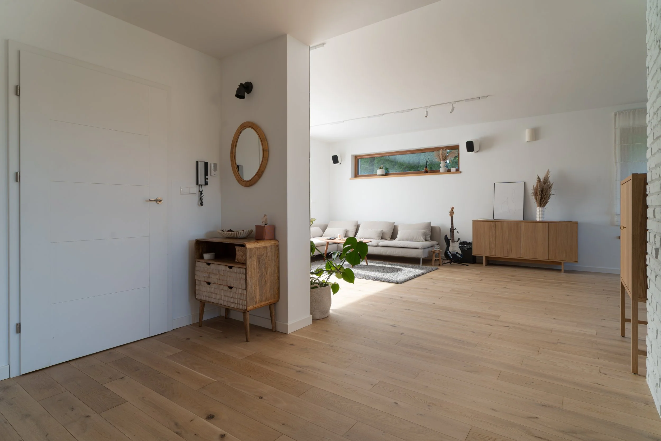 Modern living room with white walls and wooden flooring. A beige sofa set, a wooden sideboard, and a rectangular window with decorative plants. A guitar and framed art are placed against the wall, with a pot of large-leaf plant nearby.