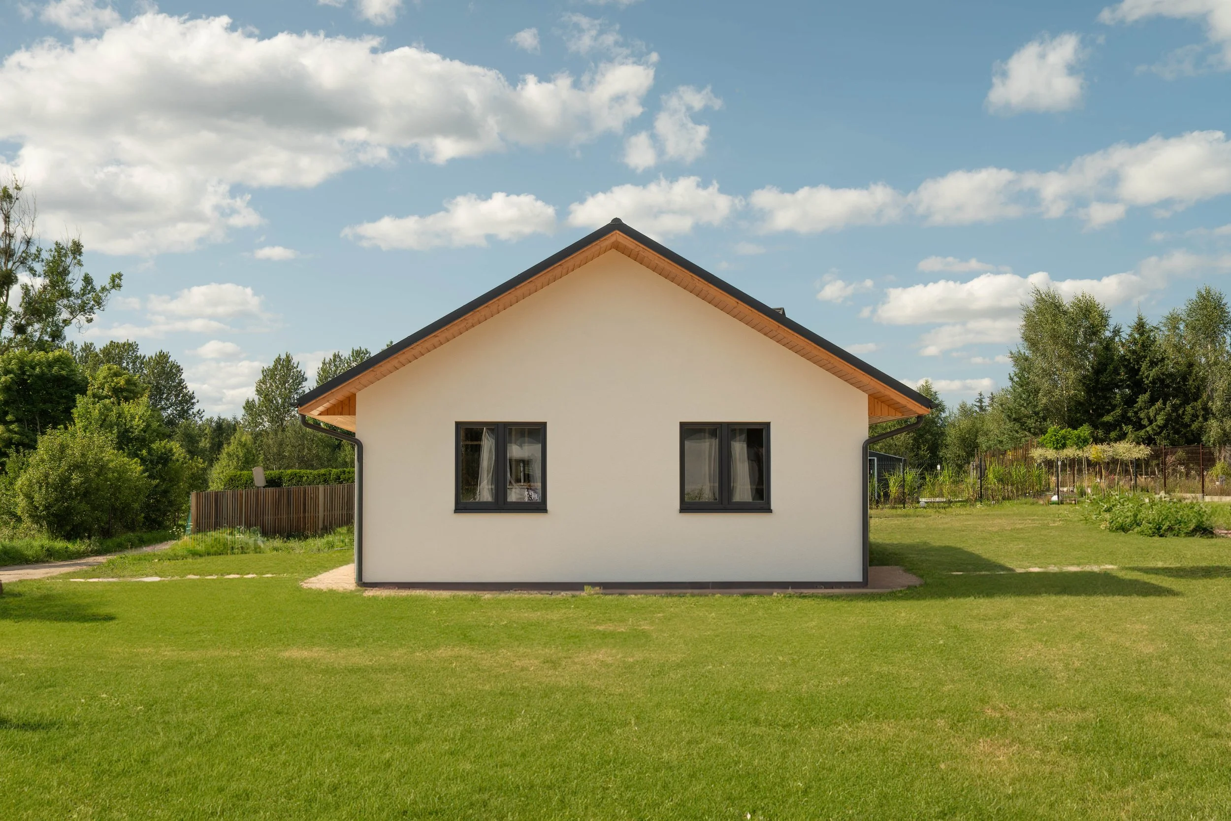A white house with black-framed windows, a sloped roof with gables, surrounded by a green lawn and trees, under a partly cloudy sky.