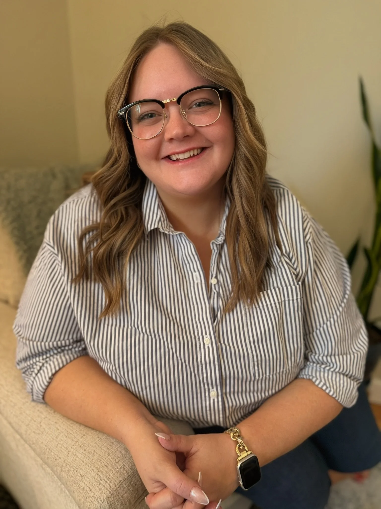 A woman with glasses smiling, wearing a striped shirt, sitting on a beige couch with a plant nearby.