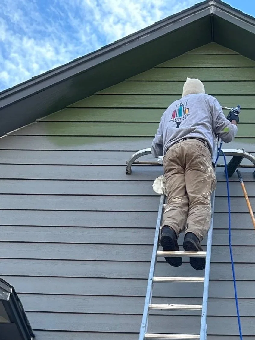 Person painting a house exterior with blue paint from a ladder, using protective coverings around windows and trim, under a blue sky.