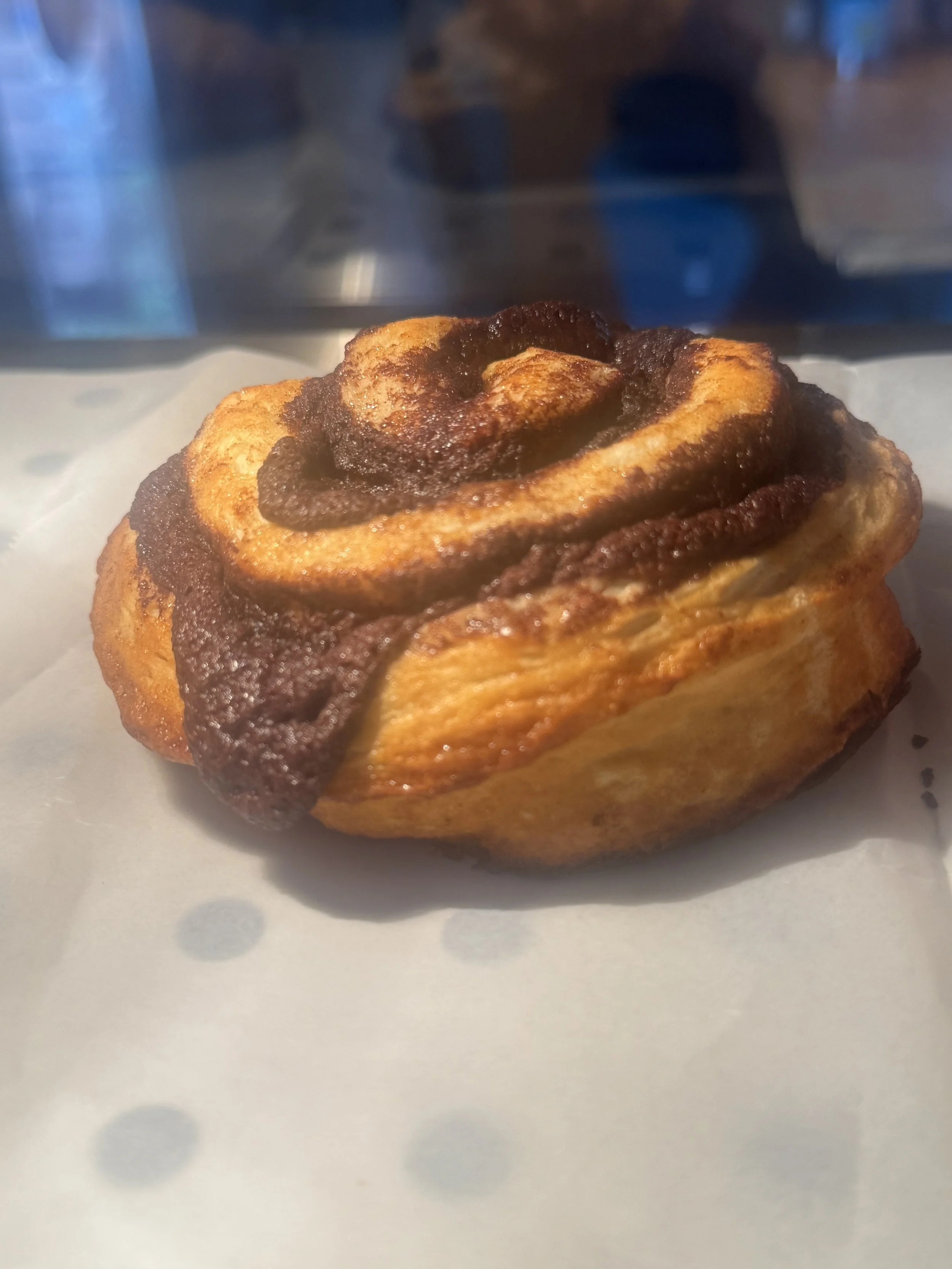 Close-up of a cinnamon roll with swirled cinnamon and sugar, golden brown on top, sitting on white parchment paper.