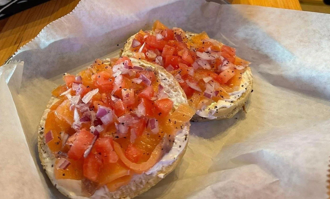 Two toasted bagel halves topped with chopped tomatoes, onions, and diced vegetables, served on parchment paper.