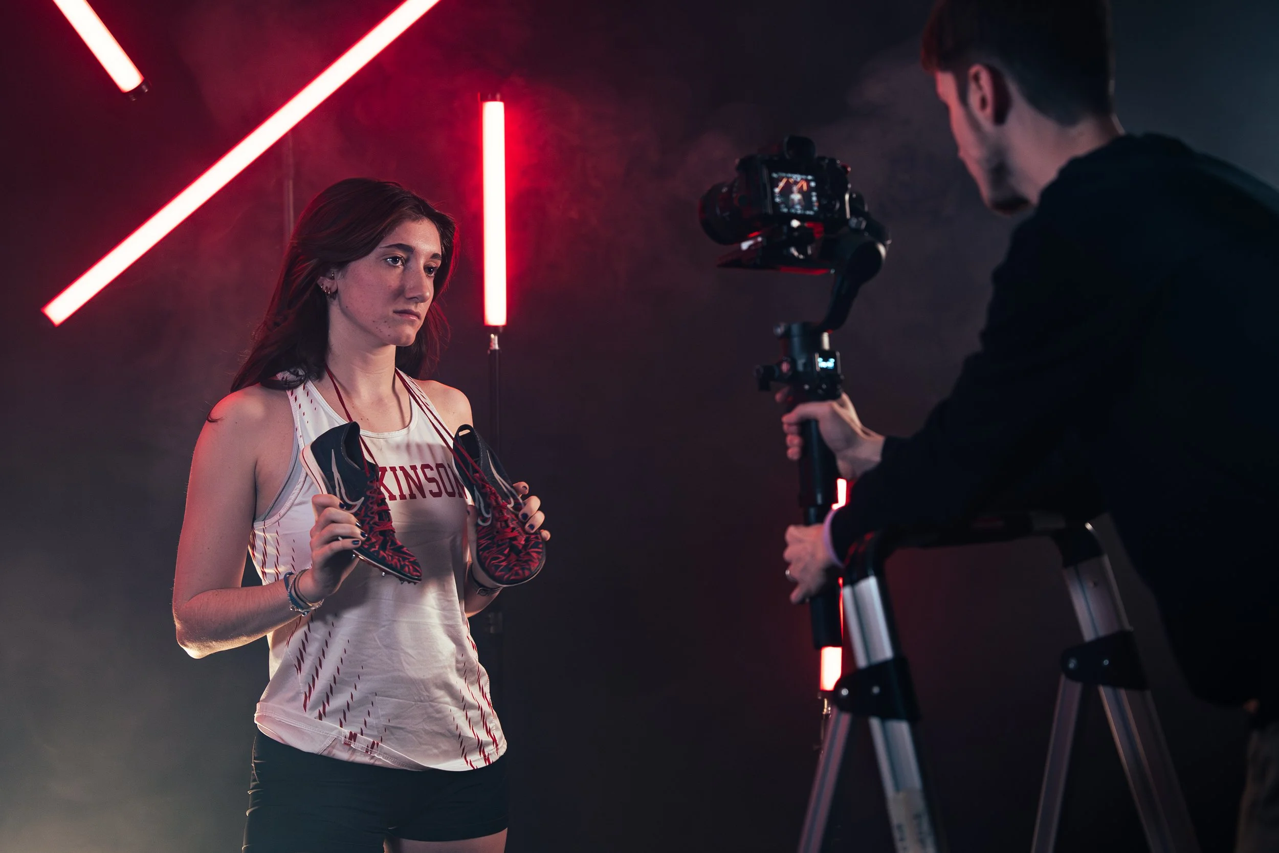 A female athlete from Dickinson College in a sporting jersey holding a pair of Nike shoes, being filmed by Tyler Caruso with a camera on a gimbal in a studio with red neon lighting.