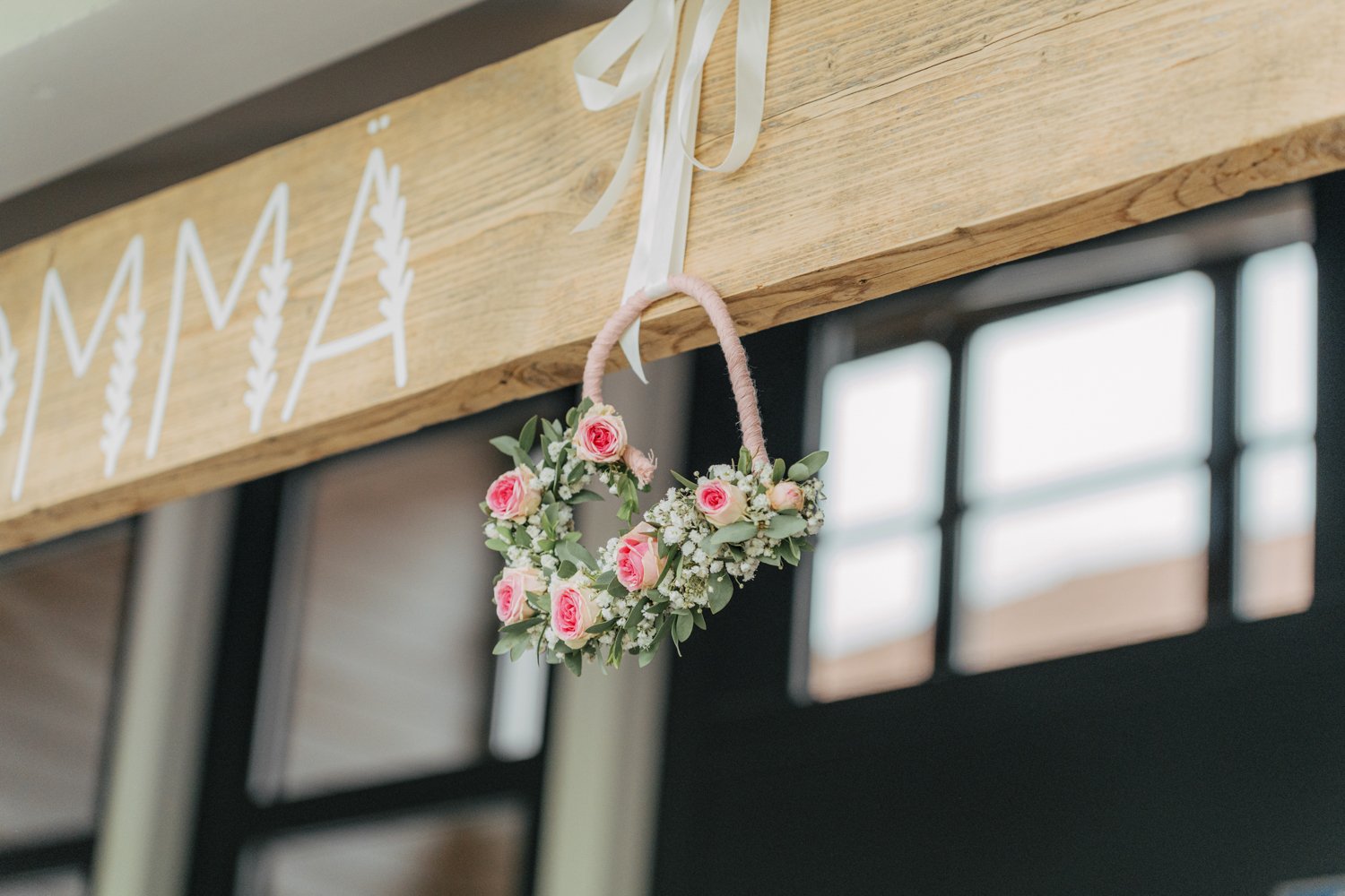 Ein Holzbalken mit weißen Buchstaben und einem Blumenkranz in Herzform, aufgehängt mit einem weißen Band.