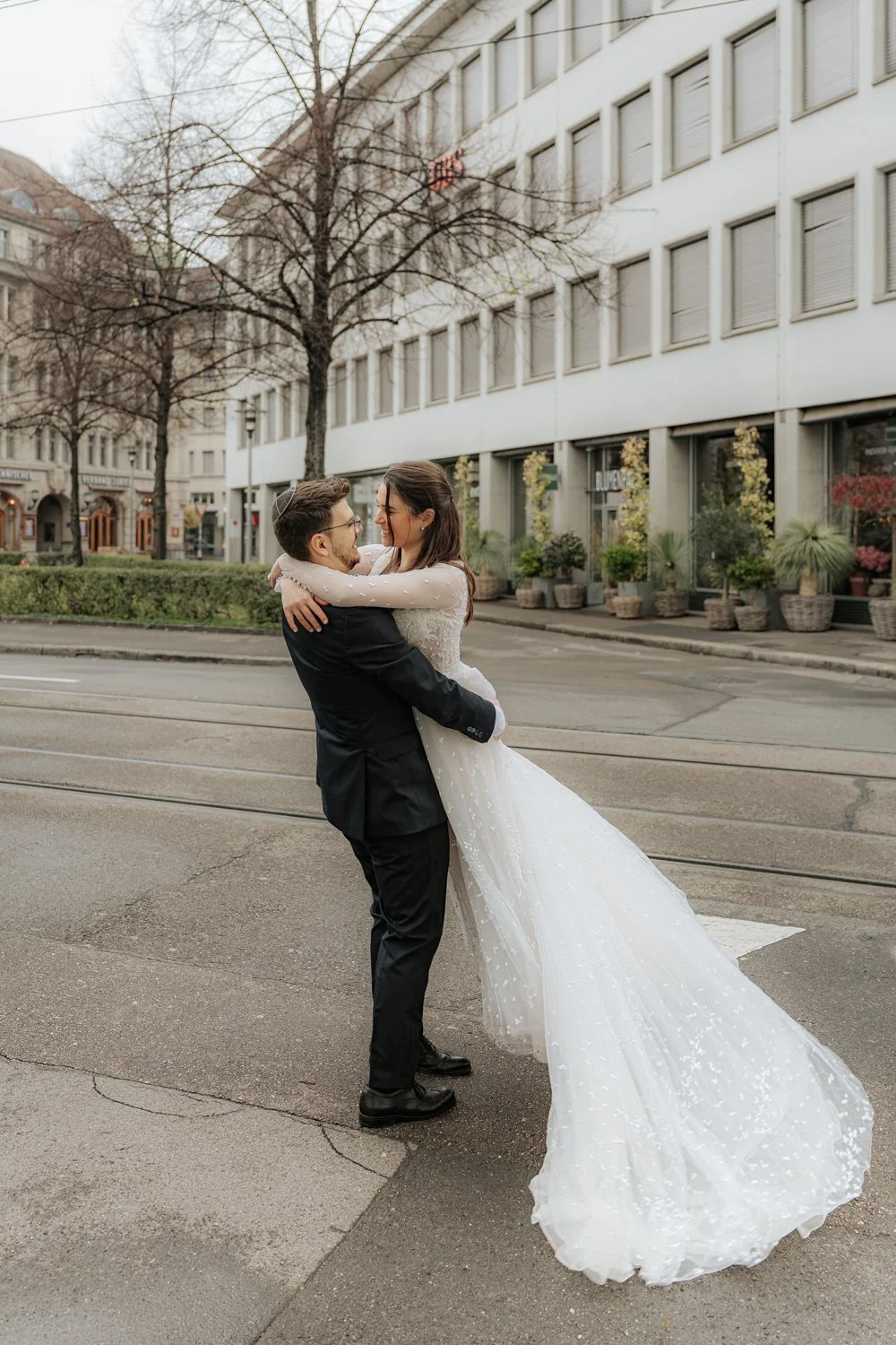 Ein lachendes Paar in Hochzeitskleidung umarmt sich auf einer Straßenkreuzung in einer Stadt, im Hintergrund Bäume und eine moderne Gebäudefassade.