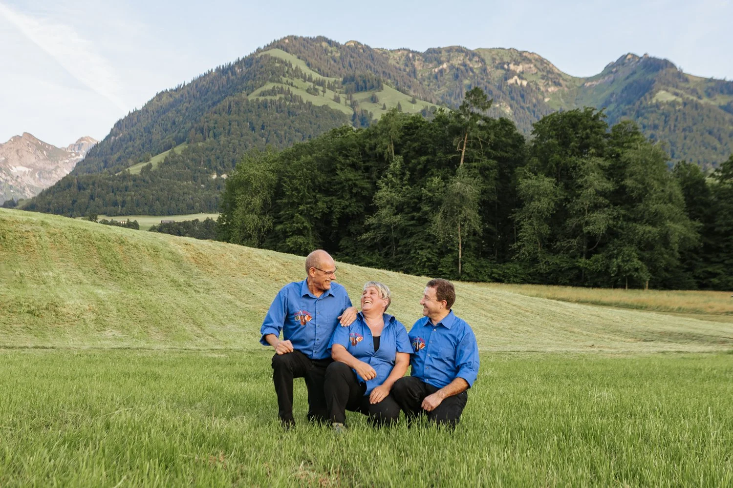 Drei Menschen in blauen Hemden sitzen lachend auf einer Wiese vor Bergen und Bäumen.