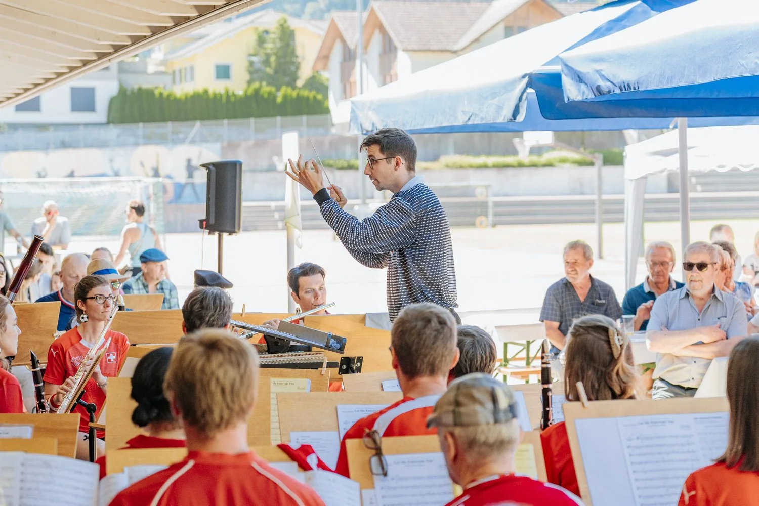 Ein Musik, geleitet von einem Dirigenten, mit einem Publikum im Hintergrund, einige mit Sonnenbrillen, bei Sonnenwetter.