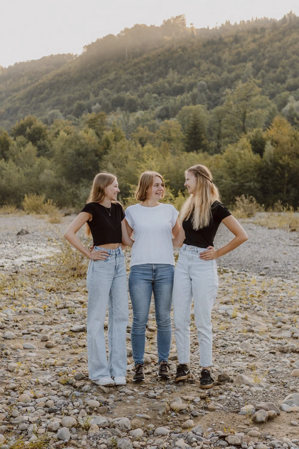 Drei junge Frauen stehen auf einem kiesbedeckten Flussbett vor einem bewaldeten Hügel und sprechen lächelnd miteinander.