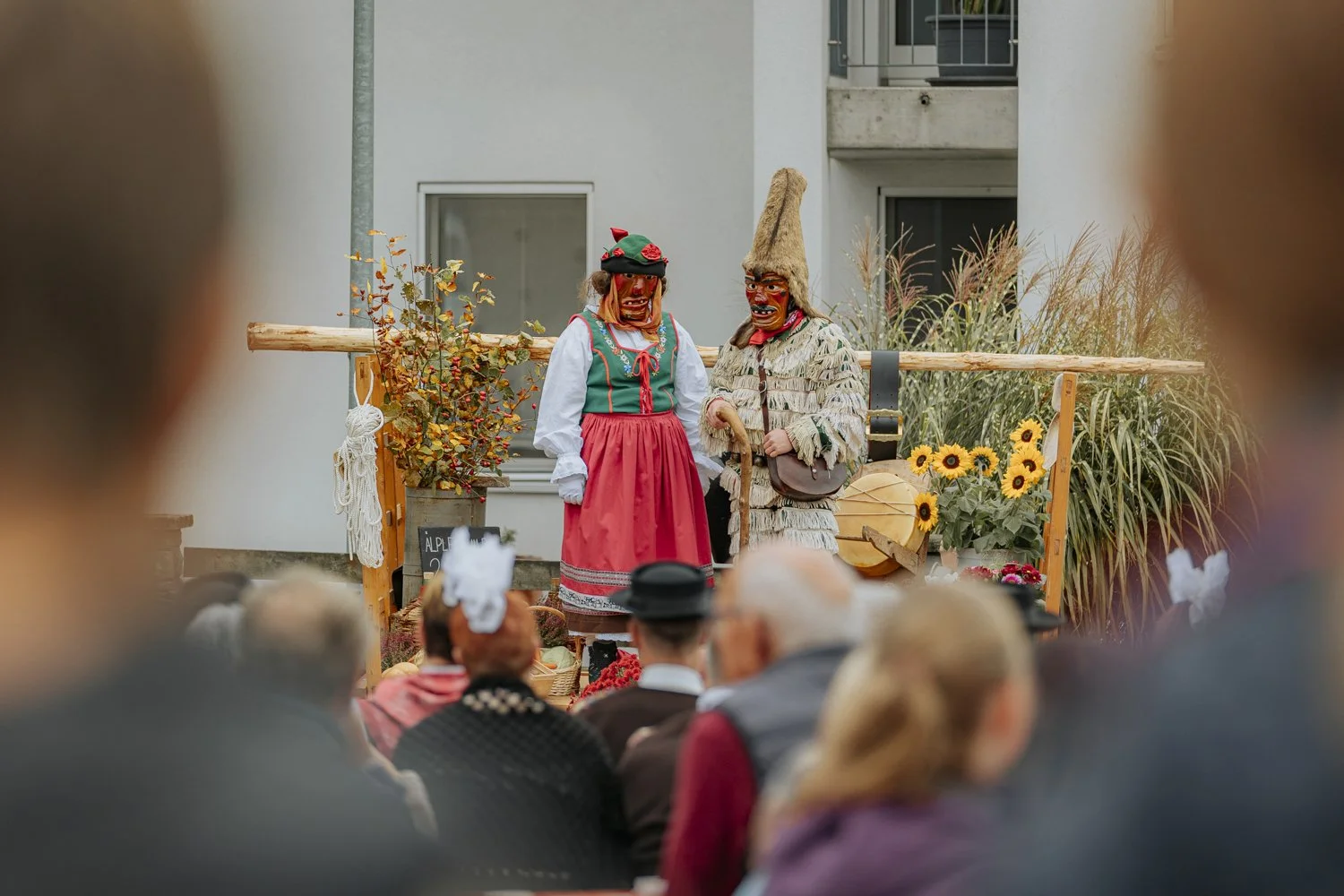 Zwei Personen in traditionellen maskierten Kostümen auf einer Bühne bei einer Veranstaltung, umgeben von Sonnenblumen und Herbstdekoration, im Hintergrund ein modernes Gebäude.