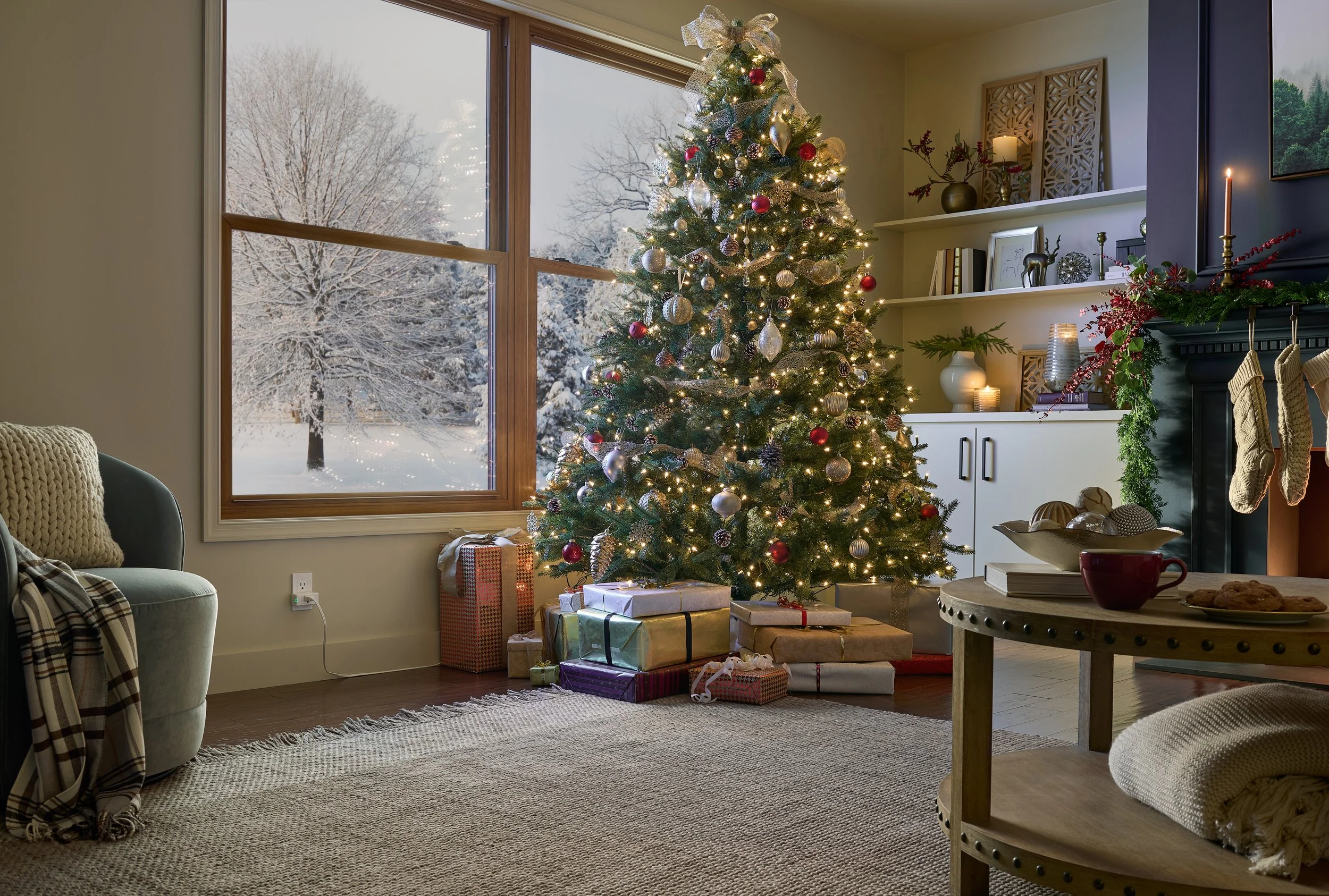 A decorated Christmas tree with lights and ornaments stands near a window with snow-covered trees outside. Wrapped presents are placed underneath the tree. The room has a cozy, decorated fireplace mantle with greenery, candles, and holiday ornaments,