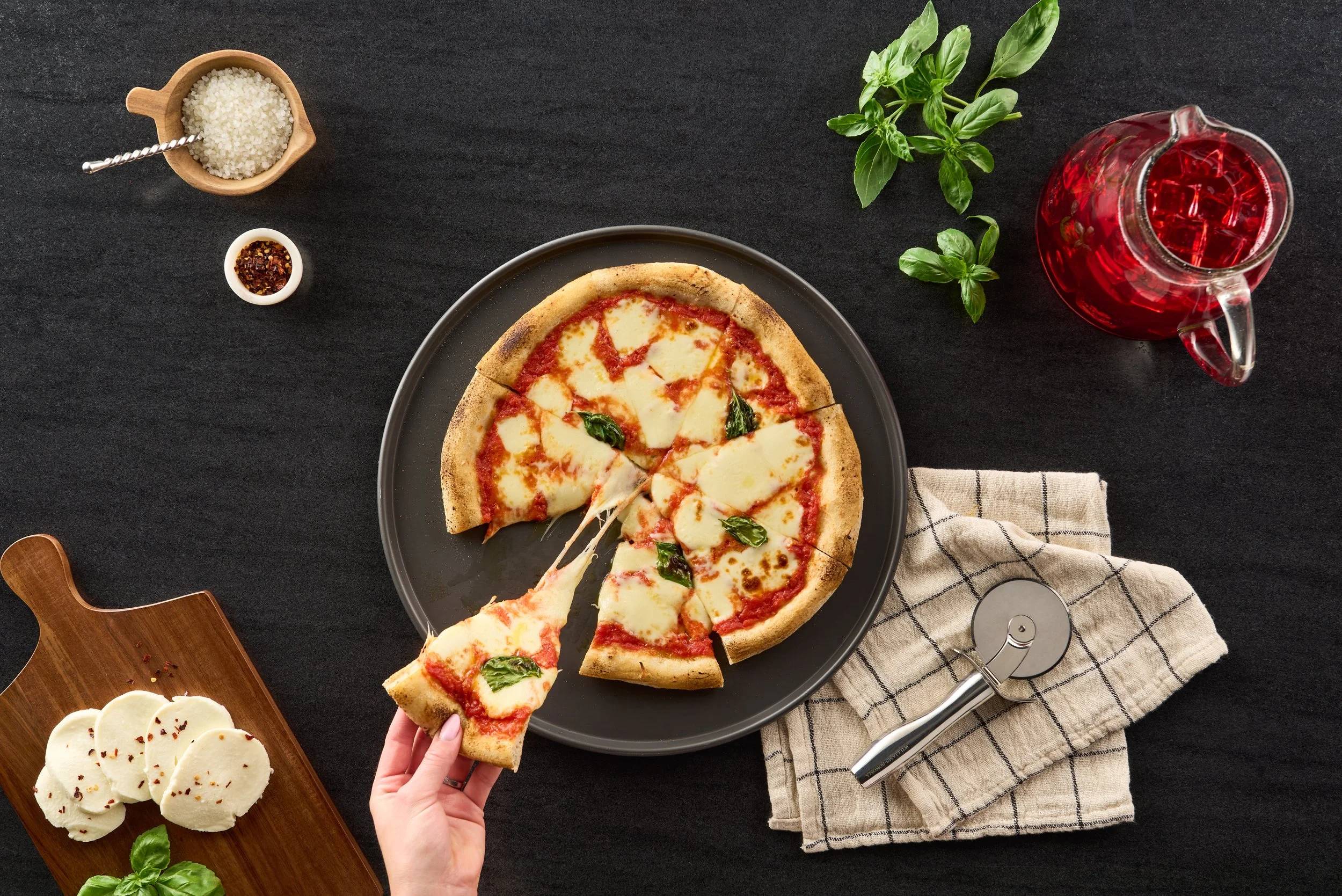 A pizza with cheese, tomato sauce, and basil on a black plate. A hand is lifting a slice of pizza. Around the pizza, there's a bunch of fresh basil, a glass jar of red drink, a small dish of red pepper flakes, a cutting board with cheese slices, and 