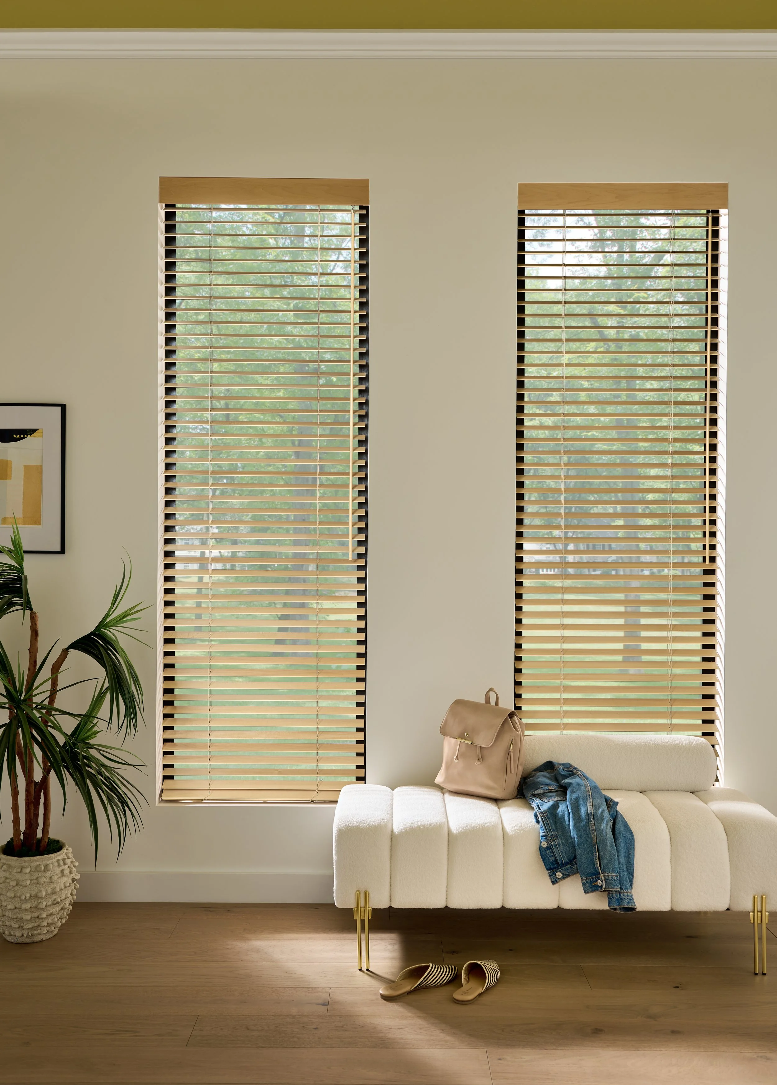 Living room with two large windows with wooden blinds, beige walls, a potted plant on the left, a framed picture, a beige sofa, a beige backpack, a pair of denim jeans, and striped slippers on the wooden floor.