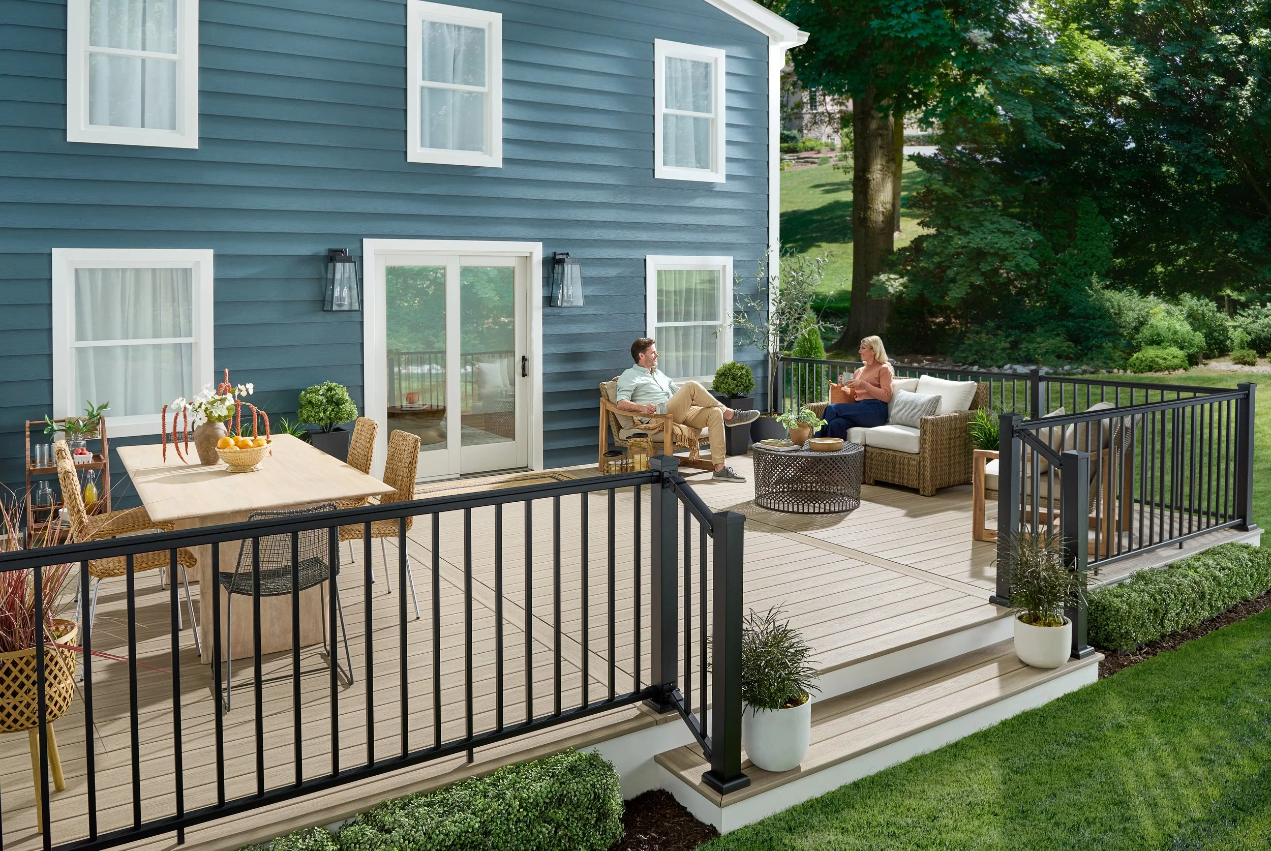 A backyard deck attached to a blue house with white trim. The deck has outdoor furniture, including a dining table with chairs and a seating area with a sofa and chairs. There are large potted plants, garden beds with greenery, and a black metal fenc