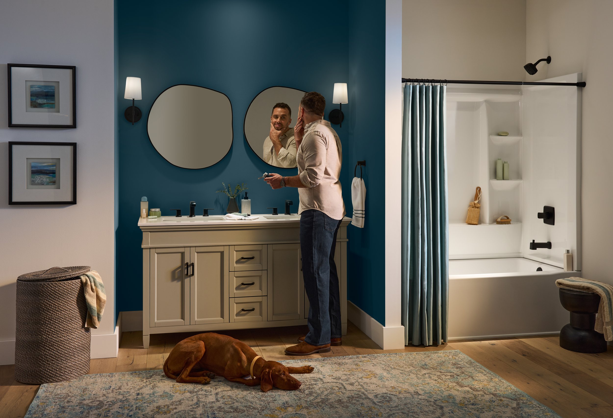 A man looks at himself in a bathroom mirror with a blue accent wall and two sconces. A brown dog lies on a colorful rug in front of him. The bathroom has a white vanity with black faucets, framed artwork on the wall, a laundry basket, and a shower wi