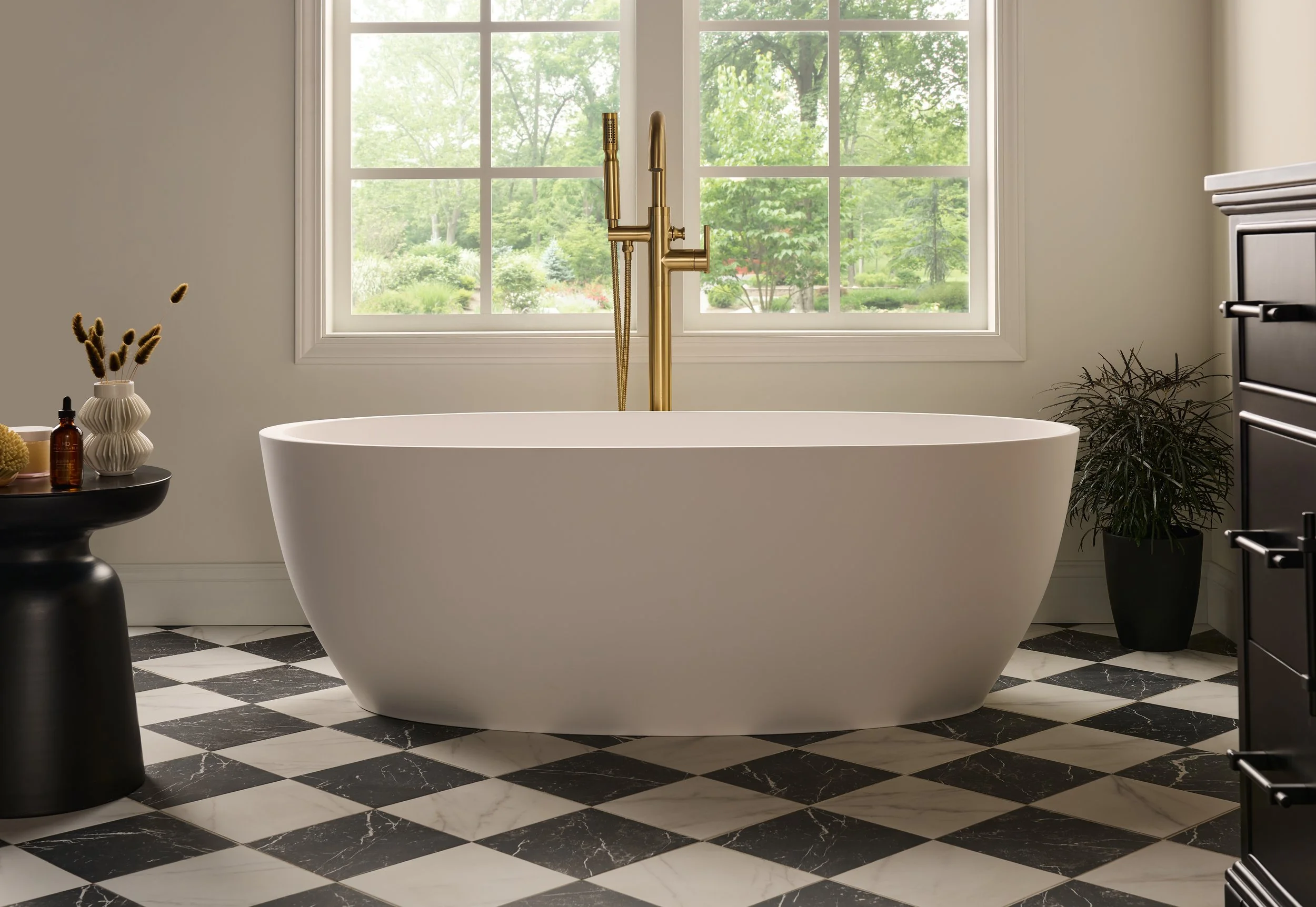 Modern bathroom with a large white bathtub, gold fixtures, a black and white checkered marble floor, a window showing greenery outside, a black side table with decor and skincare bottles, and a potted plant.