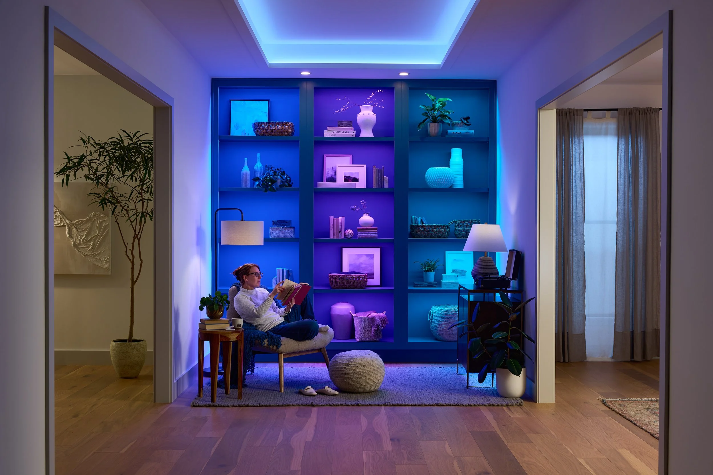 A cozy living room with a person sitting on a chair, reading a book, under a colorful blue and purple lighting display on a large bookshelf. The room has wooden flooring, a small wooden side table with books and a potted plant, a large potted plant n
