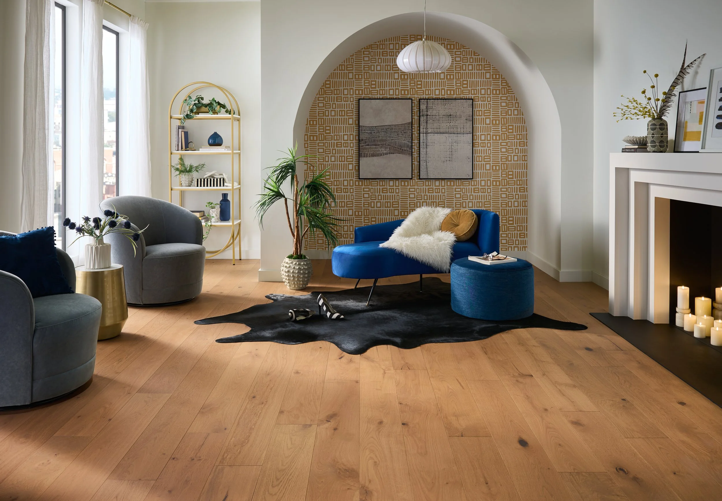 A cozy living room with hardwood floors, a black and white cowhide rug, and furniture including a gray armchair, a blue velvet sofa with fur throw, and a gray armchair. Decor includes a white vase with purple flowers, potted plants, framed artwork, c