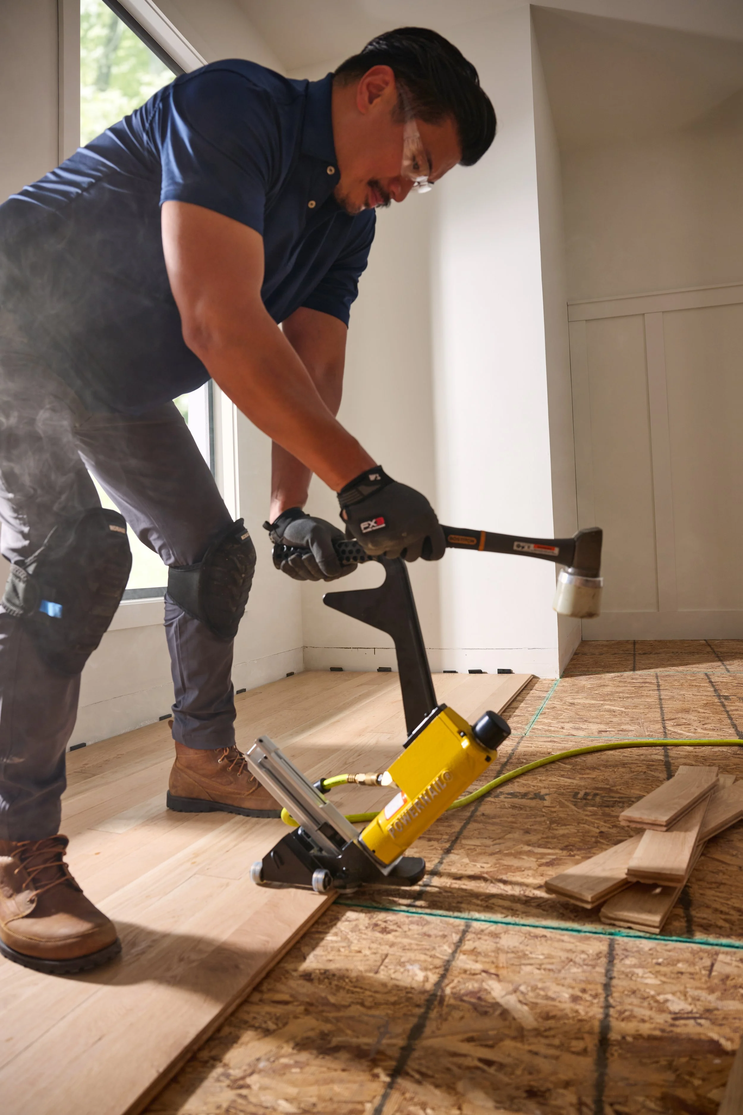 A man working on installing hardwood flooring, using a pneumatic stapler, with plywood subfloor and wooden planks around him.