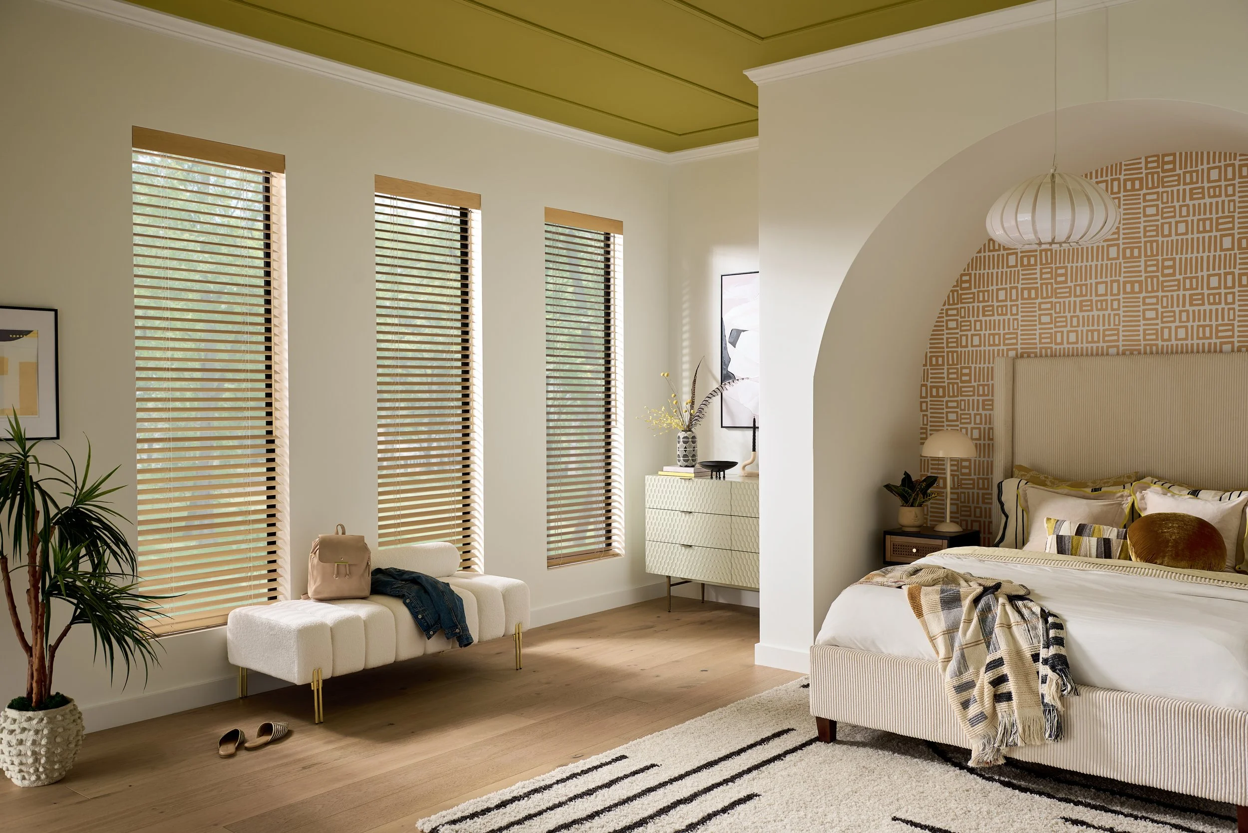 A cozy bedroom with large windows, a white sofa with a beige bag and jeans on it, a patterned wallpaper behind the bed, a hanging white pendant light, and a striped rug on wooden flooring.