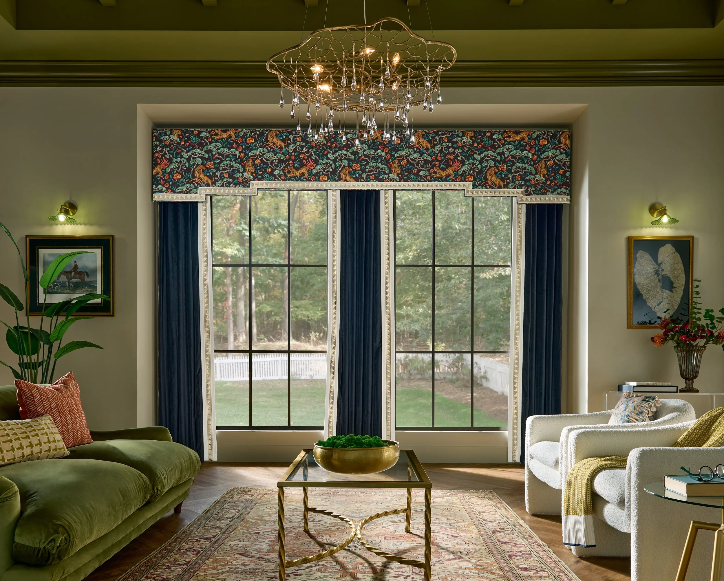 Living room with large window, green sofa with patterned pillows, white armchairs, a gold coffee table, a chandelier, framed art, and houseplants.